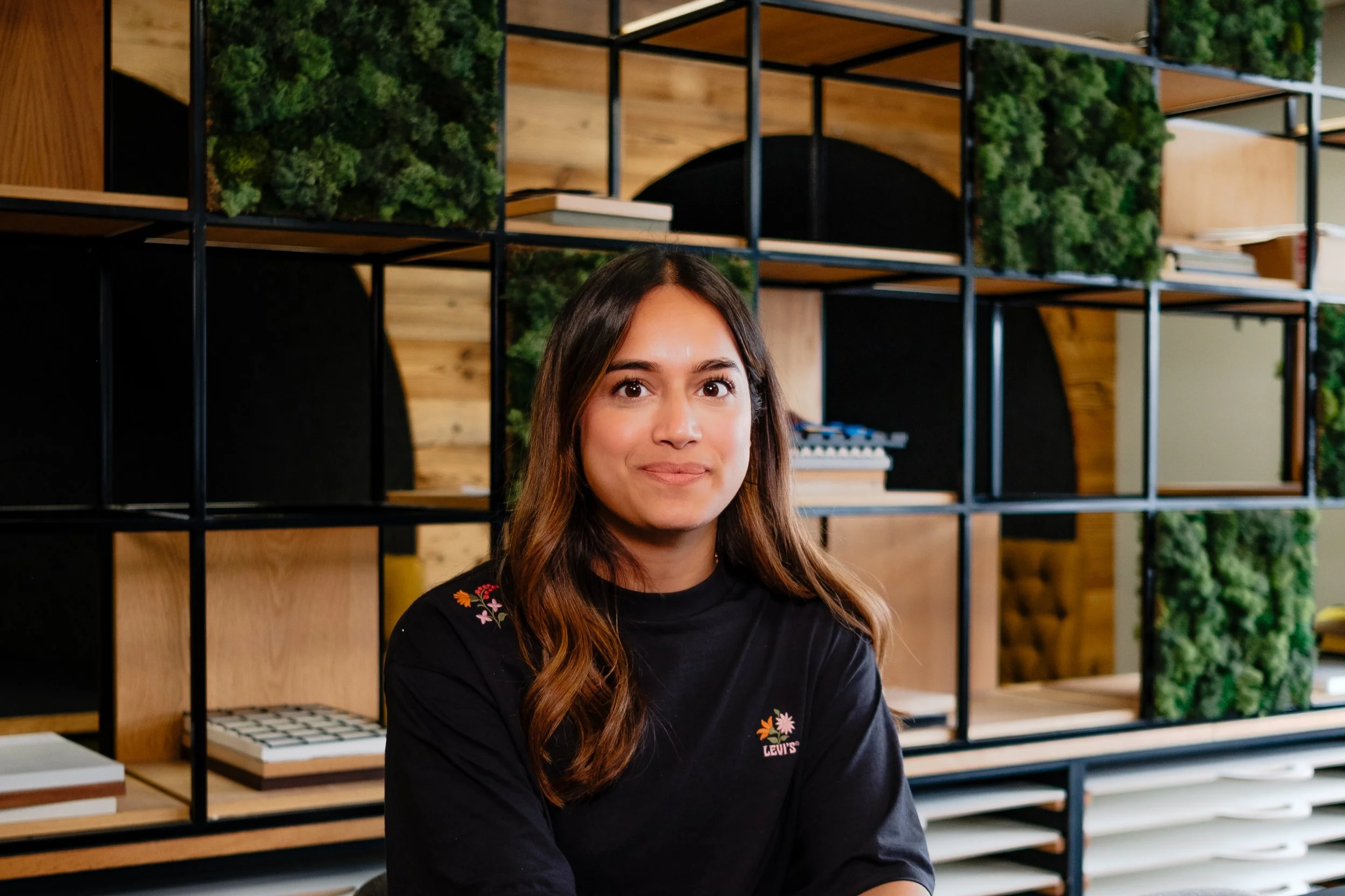 A young woman with long brown hair sitting at a black table in an office, holding a white mug, with modern shelving and green plants in the background.