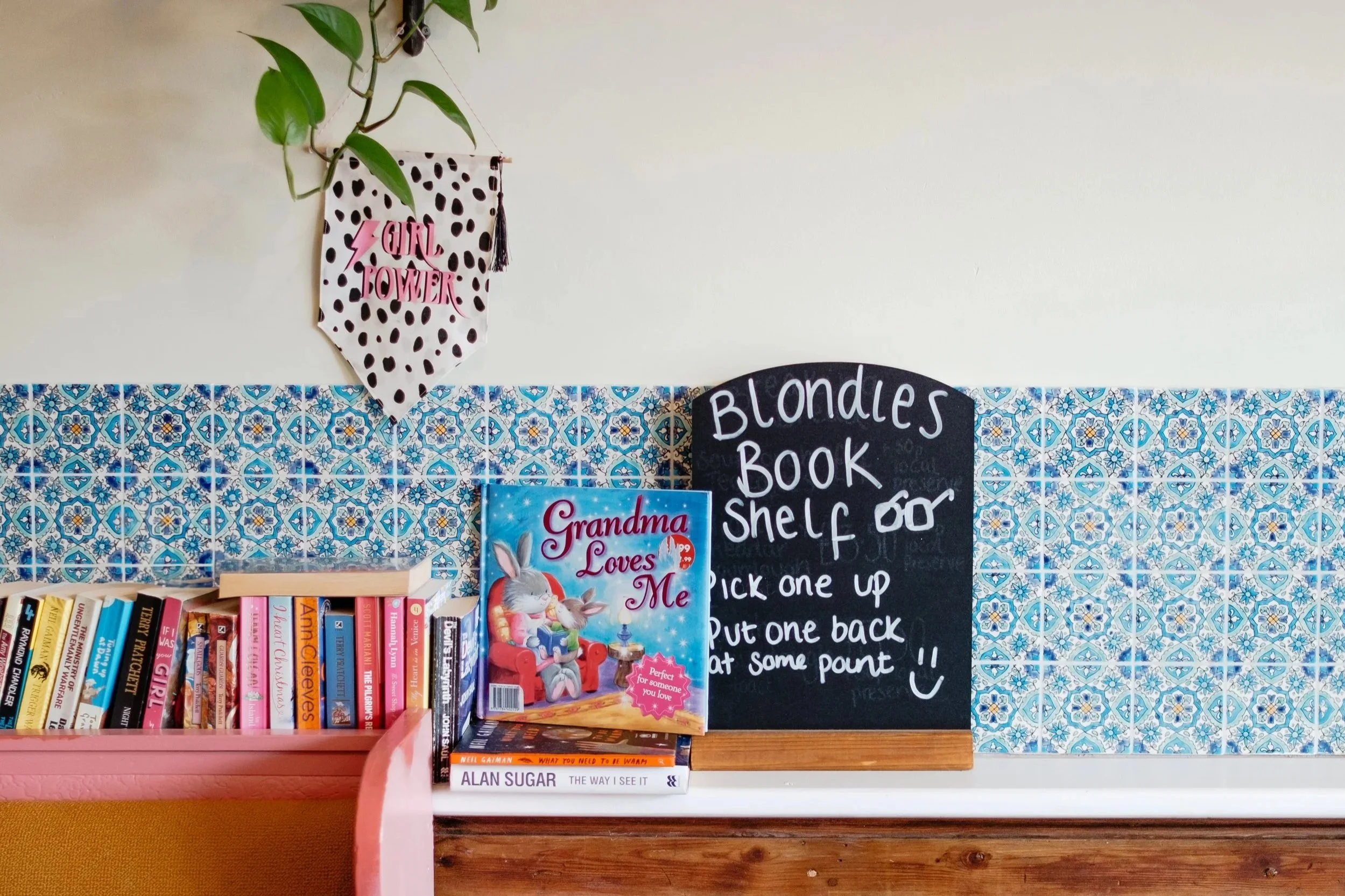 A bookshelf with colorful books next to a black chalkboard sign that says "Blondes Book Shelf" and provides instructions. A children's book titled "Grandma Loves Me" is visible on the shelf. There is a wooden chair with a light-colored seat and an or