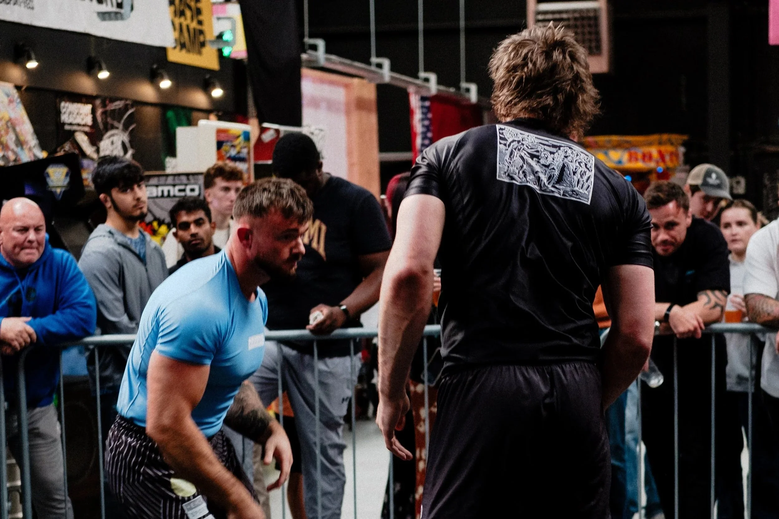 Two men barefoot on a black mat in a gym, engaged in a grappling or wrestling match, with an audience watching from behind a barrier.
