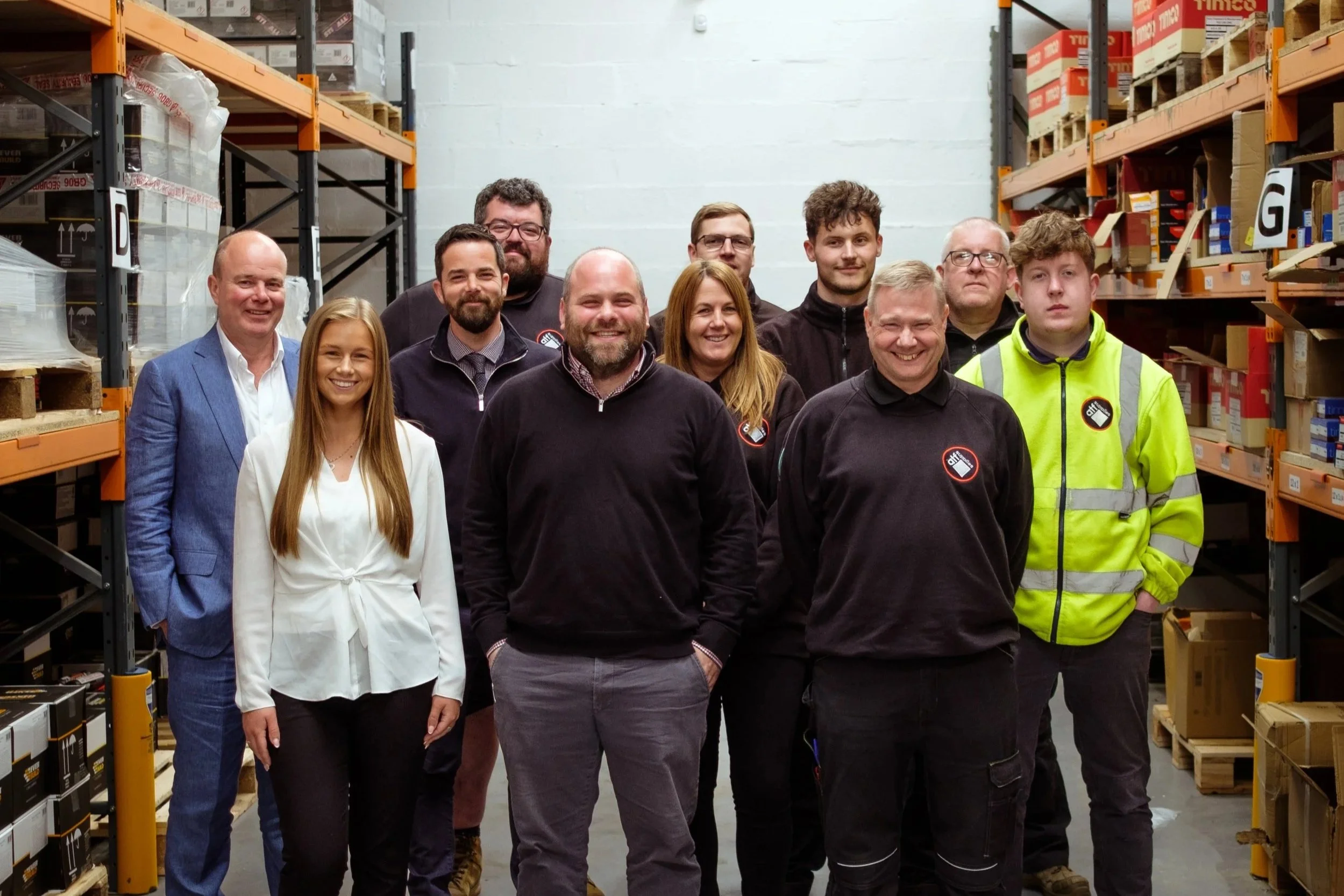 A group of people standing together in a warehouse aisle with shelves of boxes on both sides.