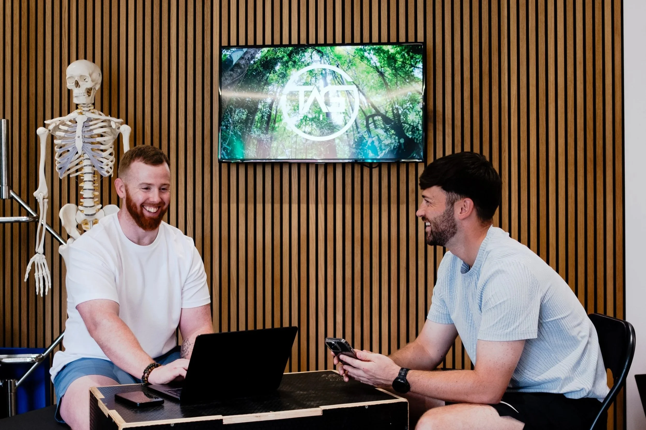 Two men sitting at a table with a laptop and a phone, laughing and talking in an office or fitness environment. A human skeleton model is visible on the left, and a screen is mounted on a wooden slat wall in the background.