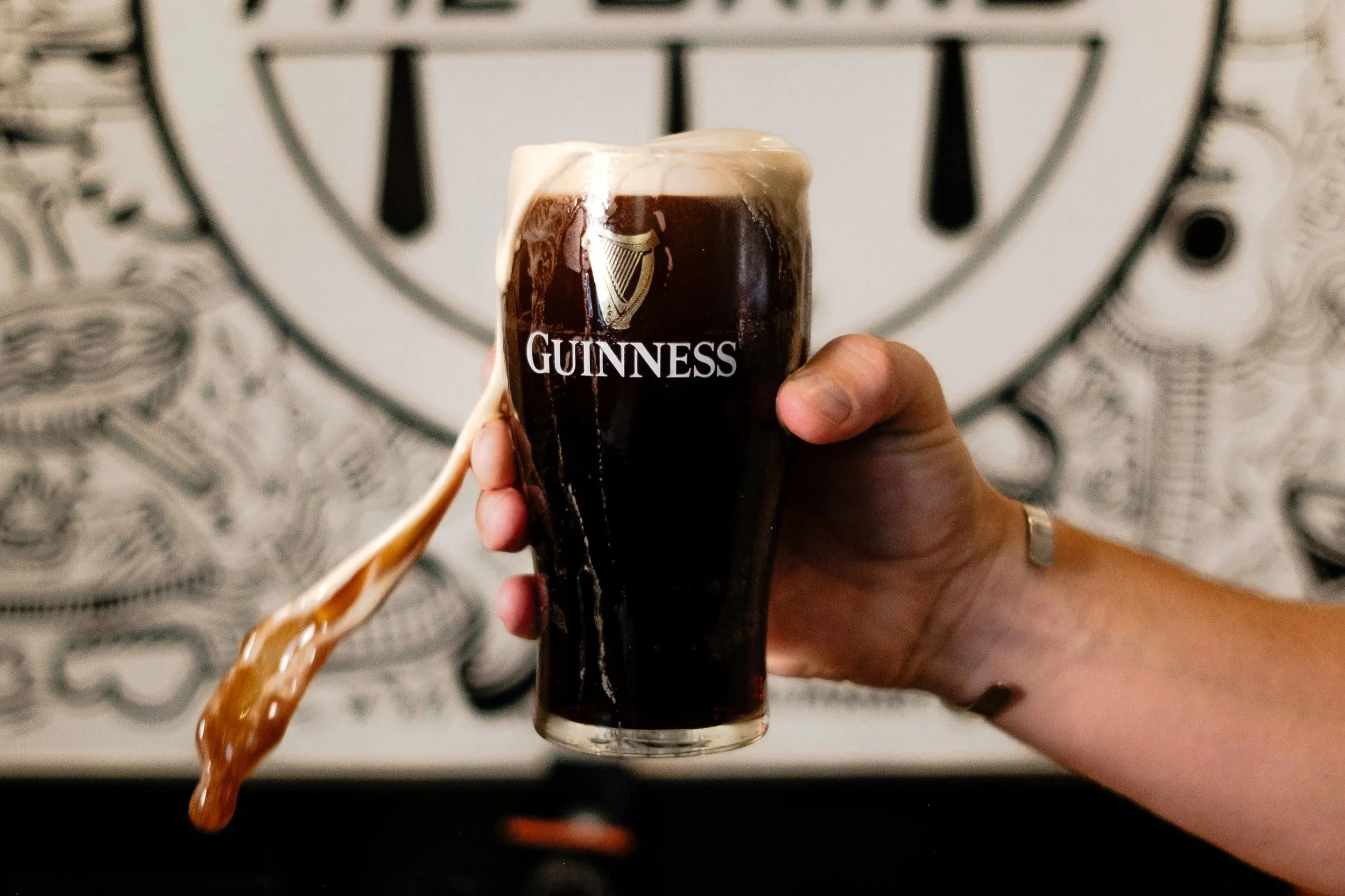 A hand holding a Guinness pint with foam overflowing, in front of a wall with a sign that reads 'The Grind'