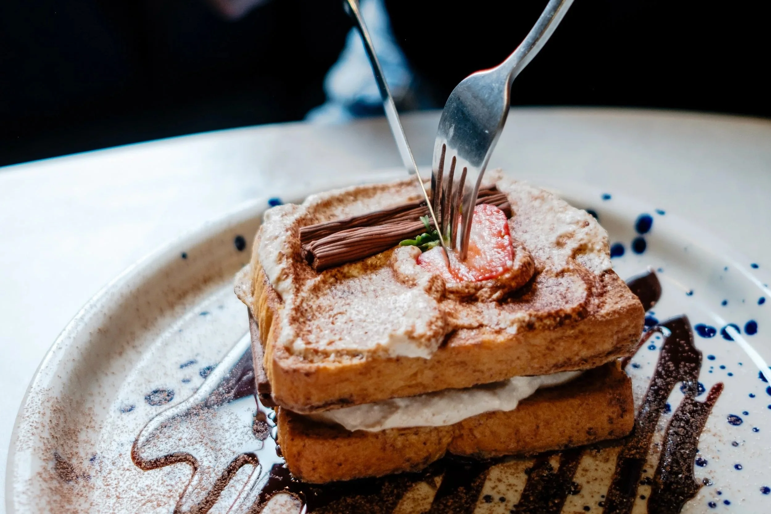 Person cutting into a layered dessert with a fork and knife, topped with chocolate and powdered sugar, on a plate decorated with chocolate drizzle.