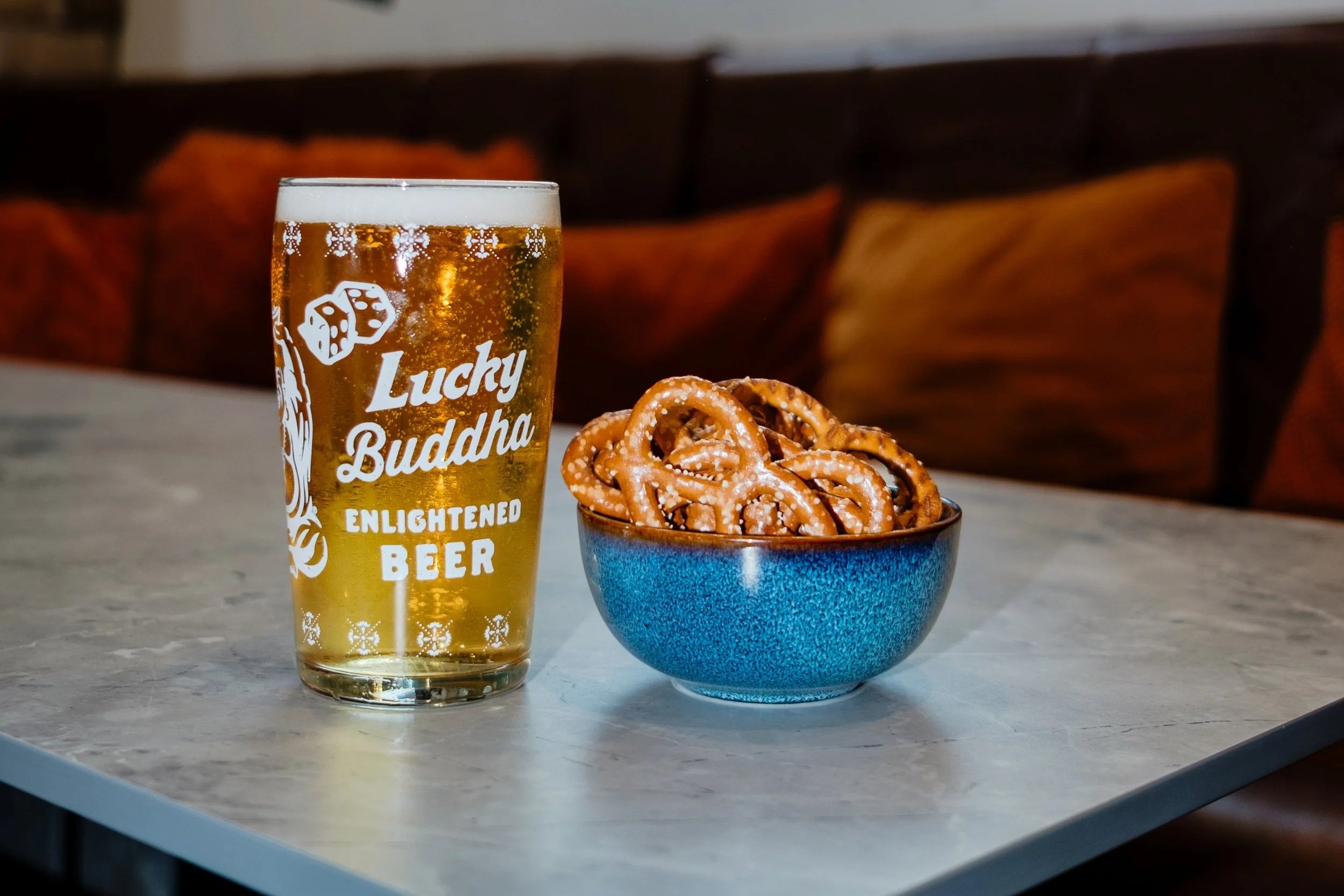 A glass of beer labeled Lucky Buddha Enlightened Beer and a bowl of pretzels on a marble table in a cozy bar or restaurant with a neon sign of a cocktail glass on the wall behind.