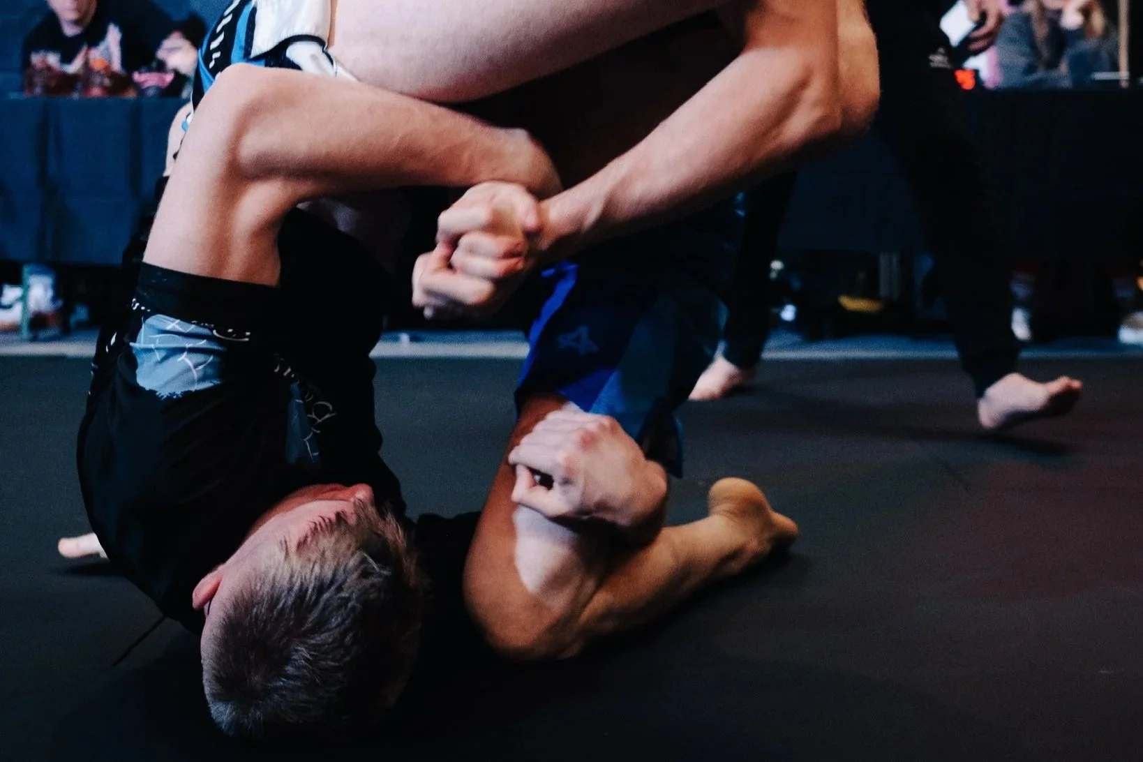 Two men competing in Brazilian Jiu-Jitsu grappling match on black mat, with spectators watching in background.