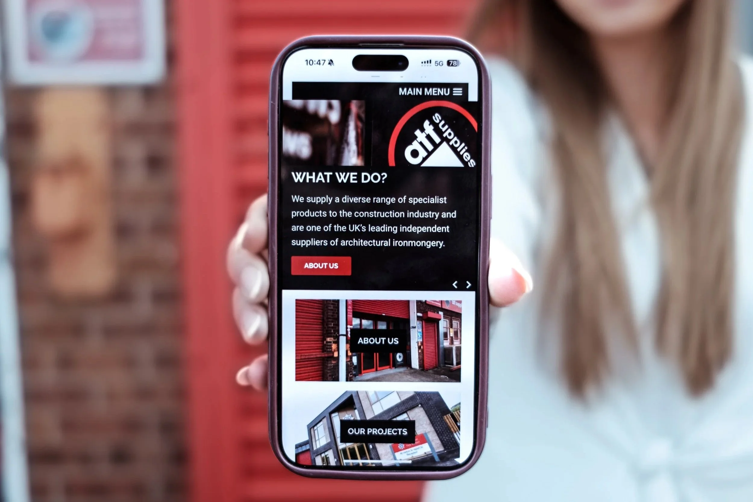 A woman holding a smartphone displaying a website for a company called 'Attrib Supplies,' with a background of a brick wall and a red paneled structure.