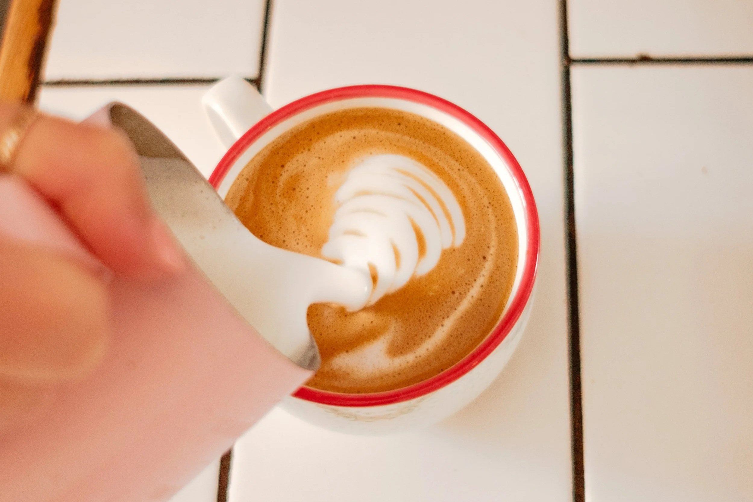 A hand pouring whipped cream into a cup of coffee with latte art, on a tiled table.