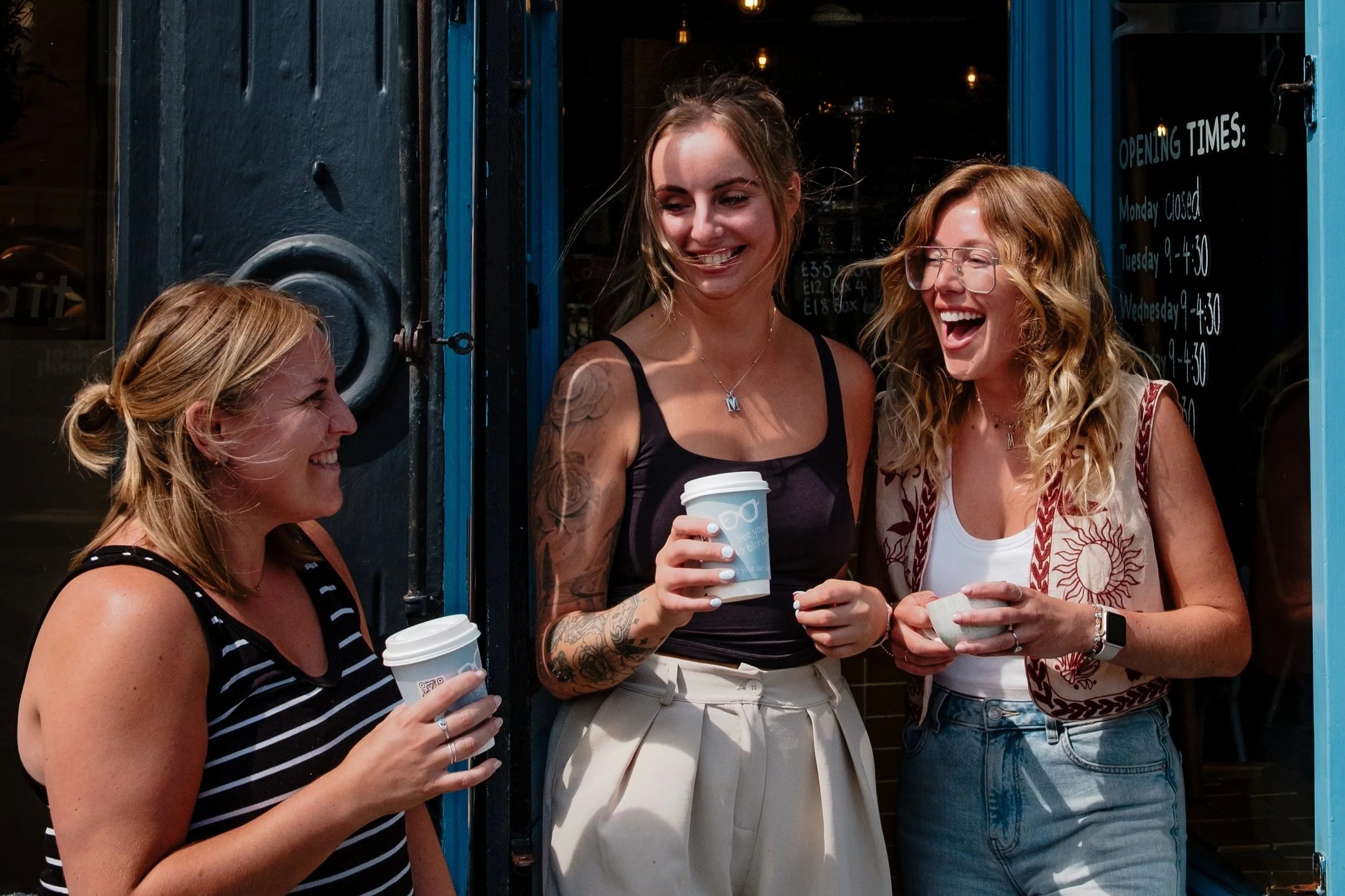 Three women standing outside a shop, smiling and holding coffee cups, engaging in conversation.