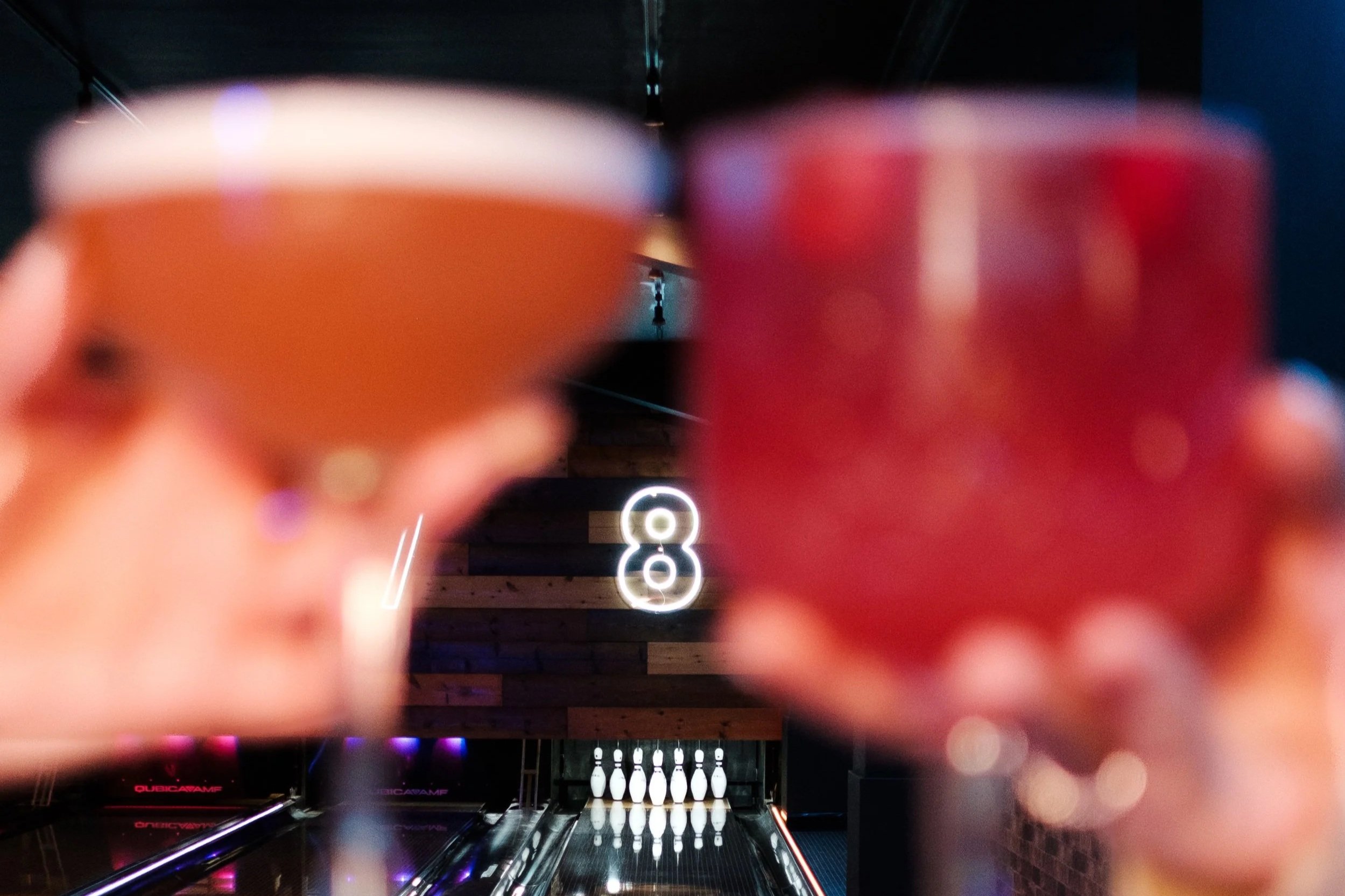 Two cocktails held up in front of a bowling alley with pins and a glowing number 8 in the background.