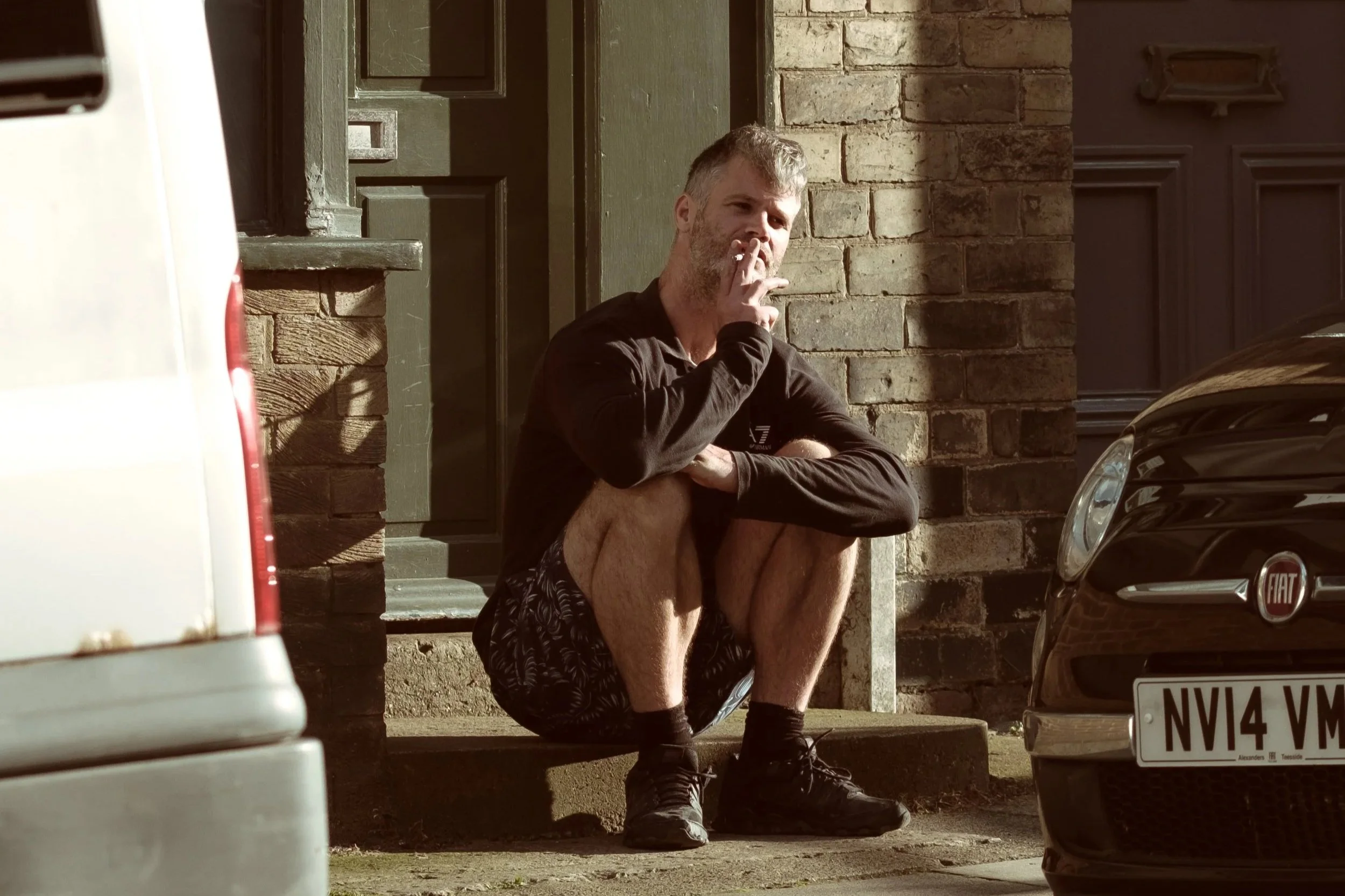 A man is squatting on the sidewalk in front of a brick building, smoking a cigarette. He is wearing a black long-sleeve shirt, patterned shorts, and running shoes. Cars are parked nearby, with the front of a gray car visible on the right and part of 