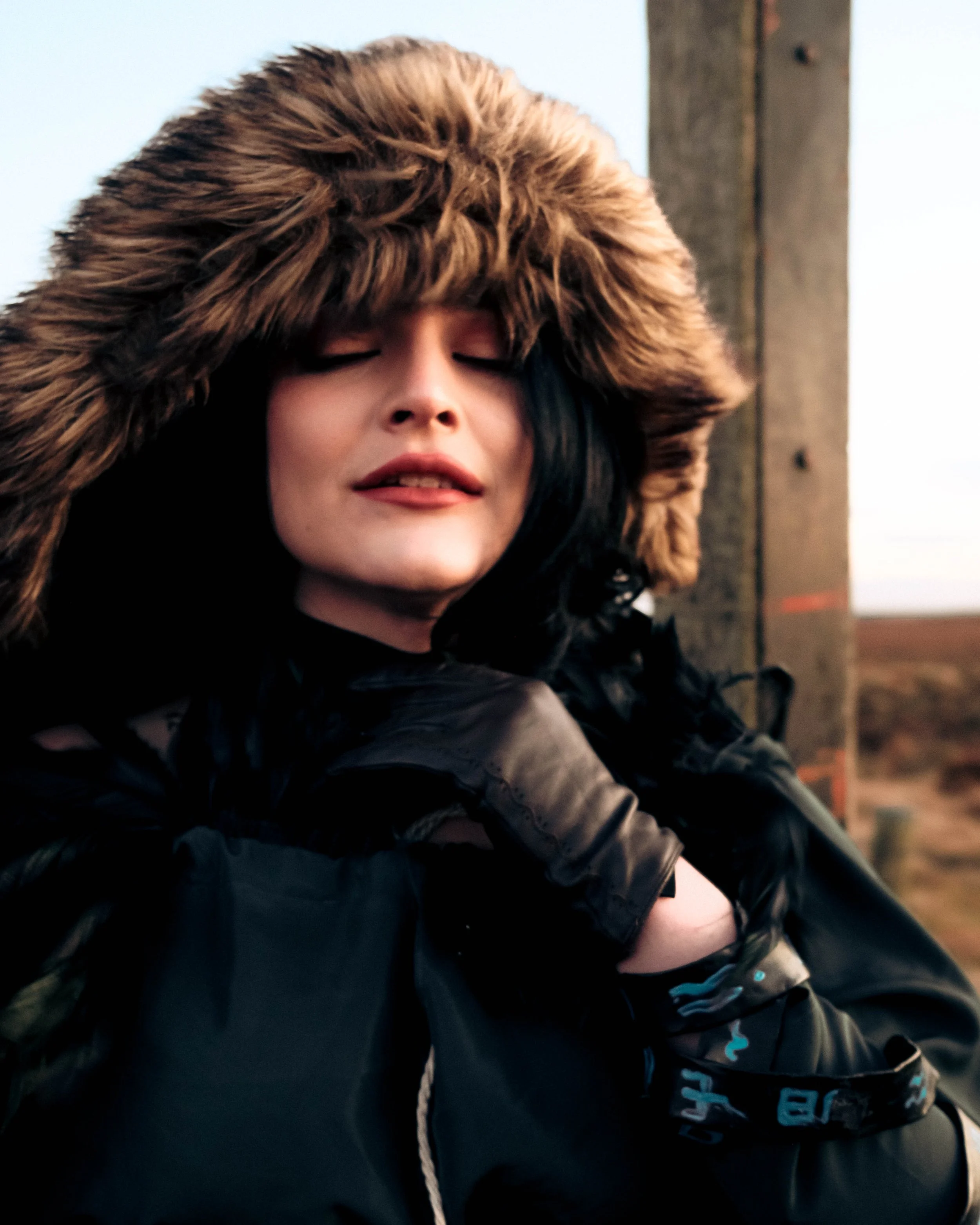 A person with dark hair wearing a large brown faux fur hood with a leather jacket, standing outdoors during sunset with a wooden fence in the background.