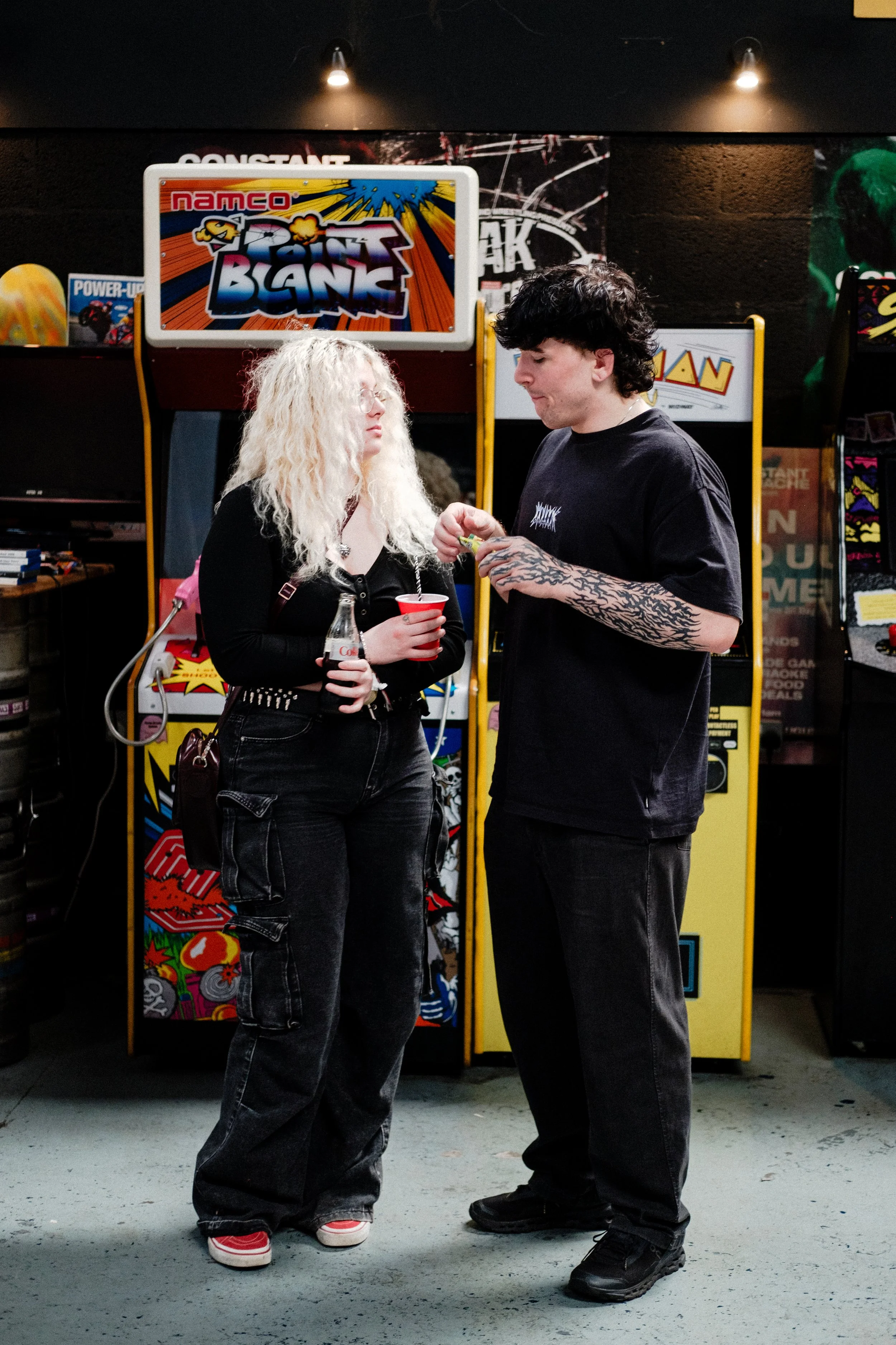 A young woman and a young man are standing face to face in front of arcade machines, engaging in conversation. The woman has long, curly blond hair, glasses, and is holding a red cup and a bottle of Coca-Cola. The man has black, wavy hair, tattoos on