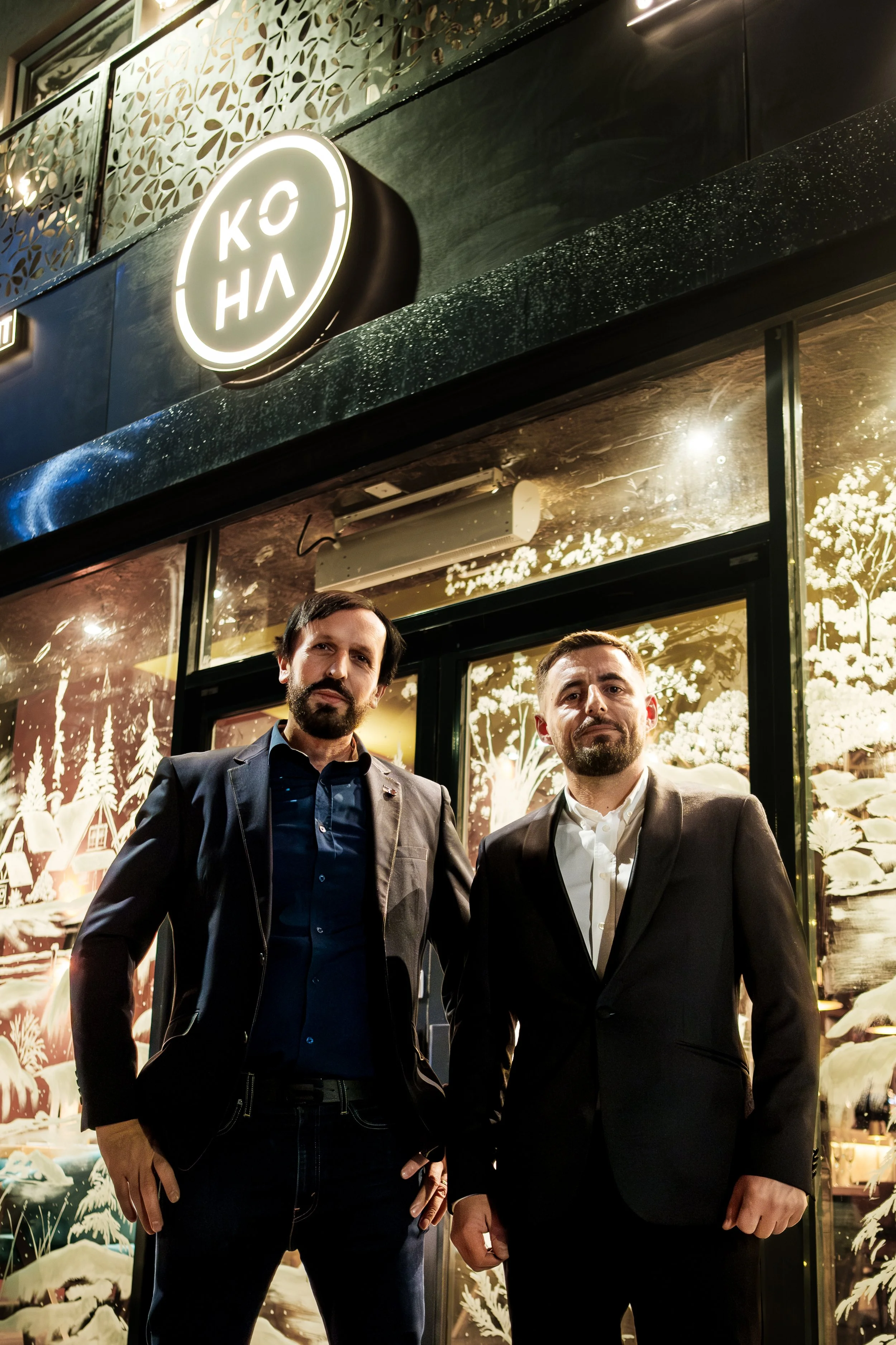 Two men in suits standing outside a restaurant named KOYO at night, with decorative illuminated windows in the background.