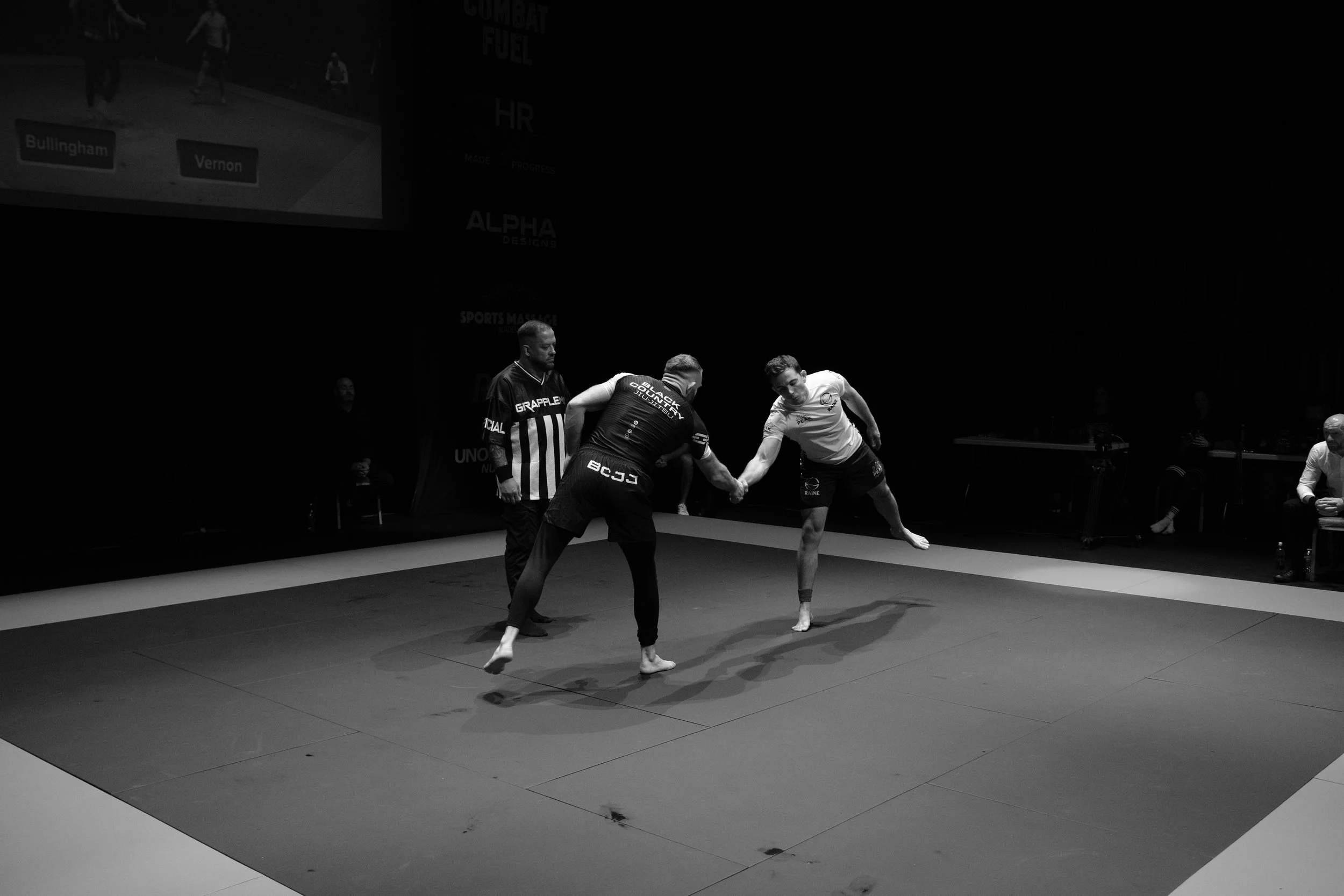 Two competitors engaged in a grappling match on a mat, with a referee and an umpire observing, in a martial arts or wrestling event.