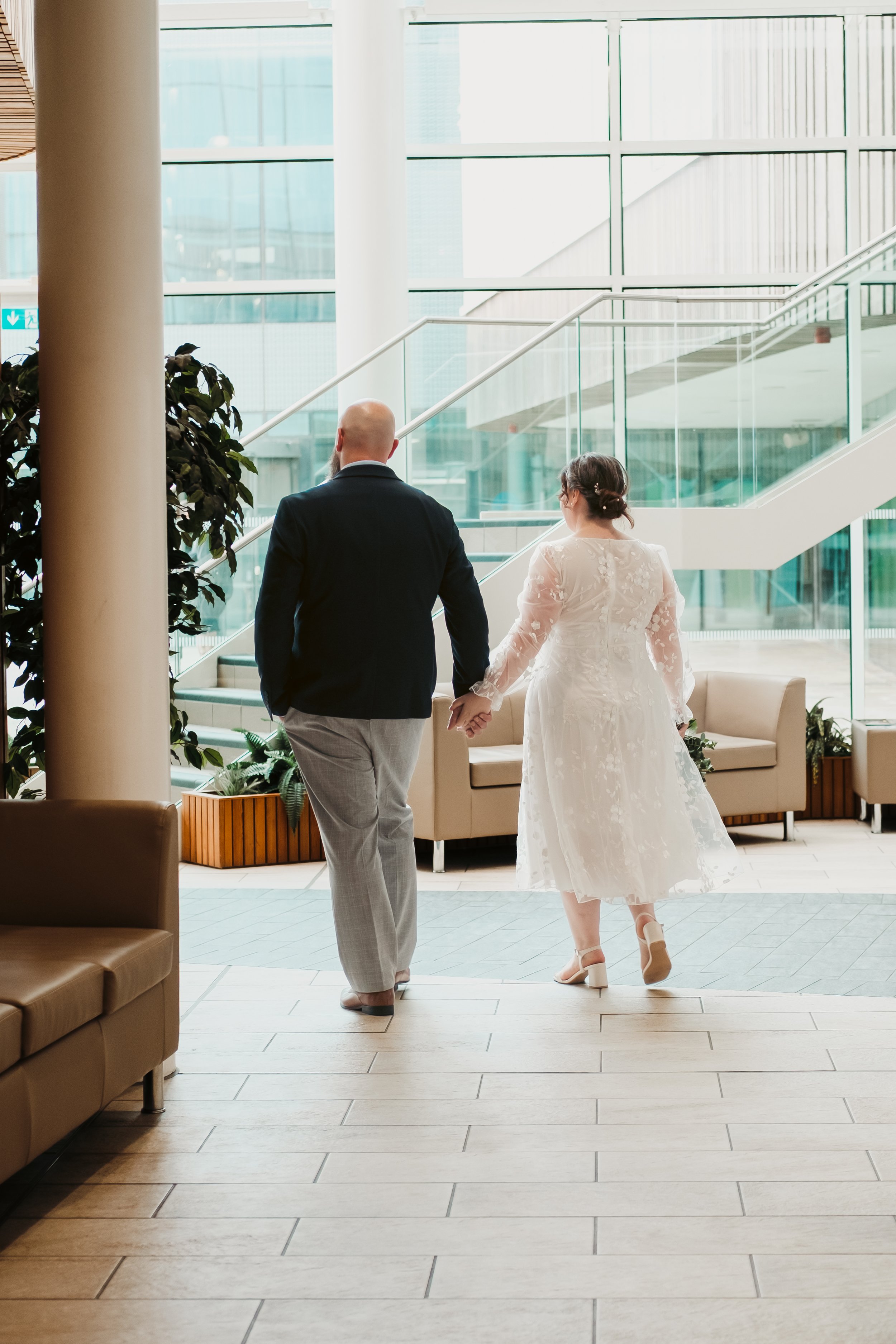 A man and woman walking hand in hand in a modern indoor space with large windows, seating, and plants, possibly at a wedding or special event.