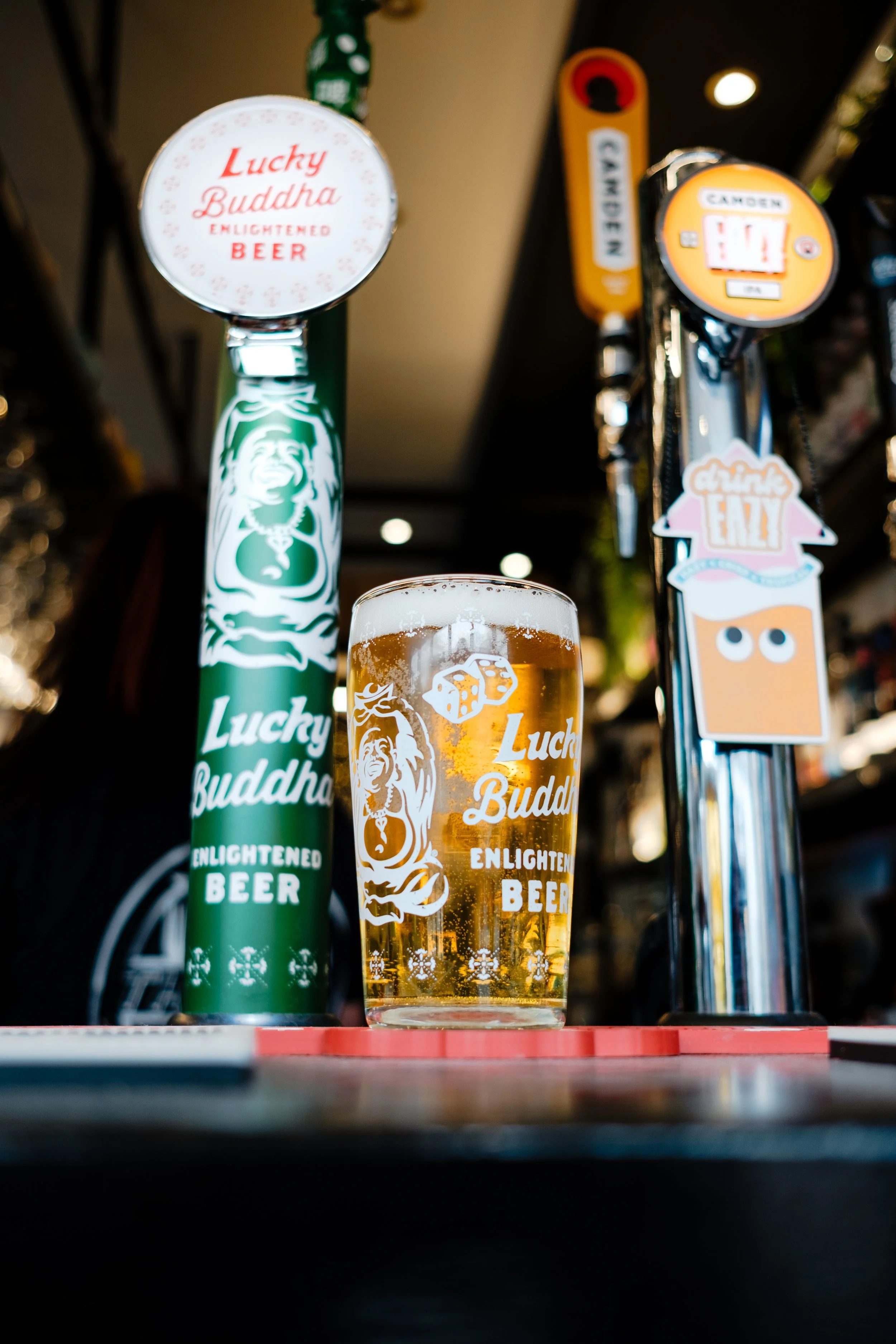 A glass of beer on a bar with a Lucky Buddha beer tap and other beer taps in the background.