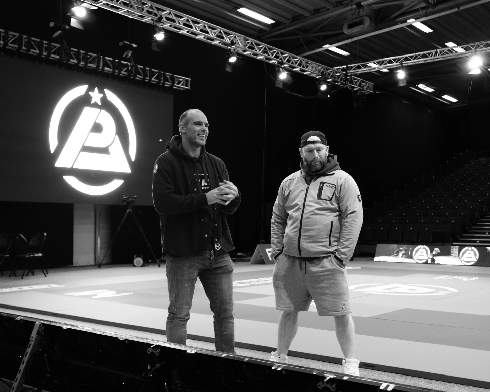 Two men standing on a martial arts mat during an event, with a large illuminated logo on a screen behind them featuring a stylized 'A' with a star above it.