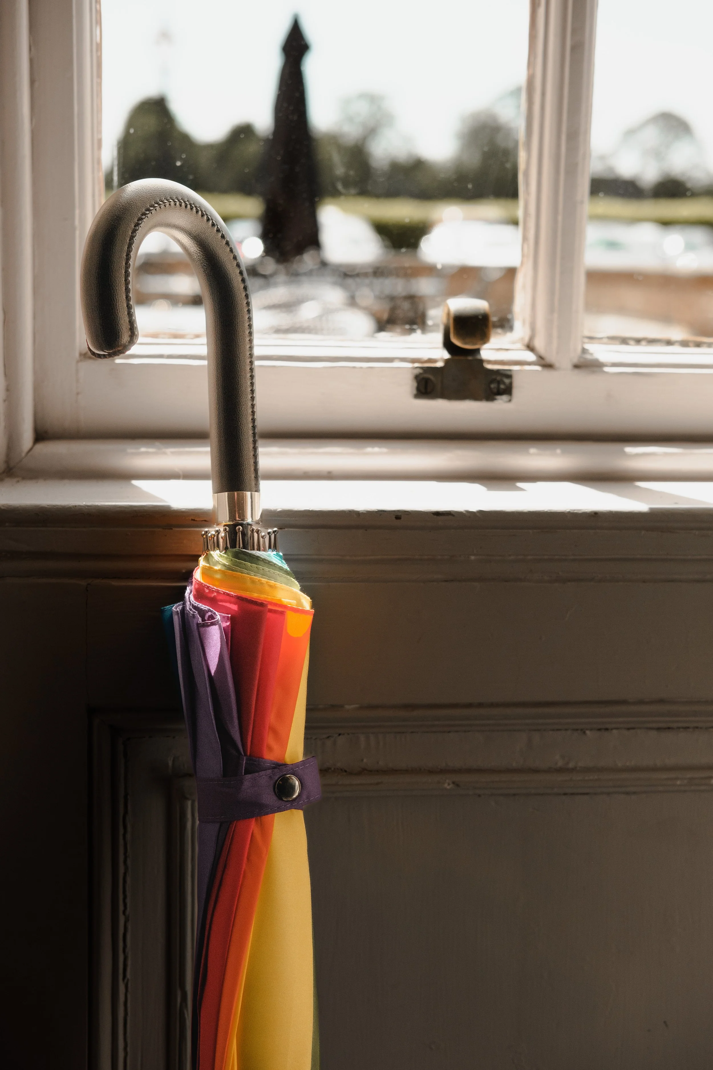 A rainbow-colored umbrella hanging from a kitchen window handle with a cloudy outdoor scene and trees visible in the background.