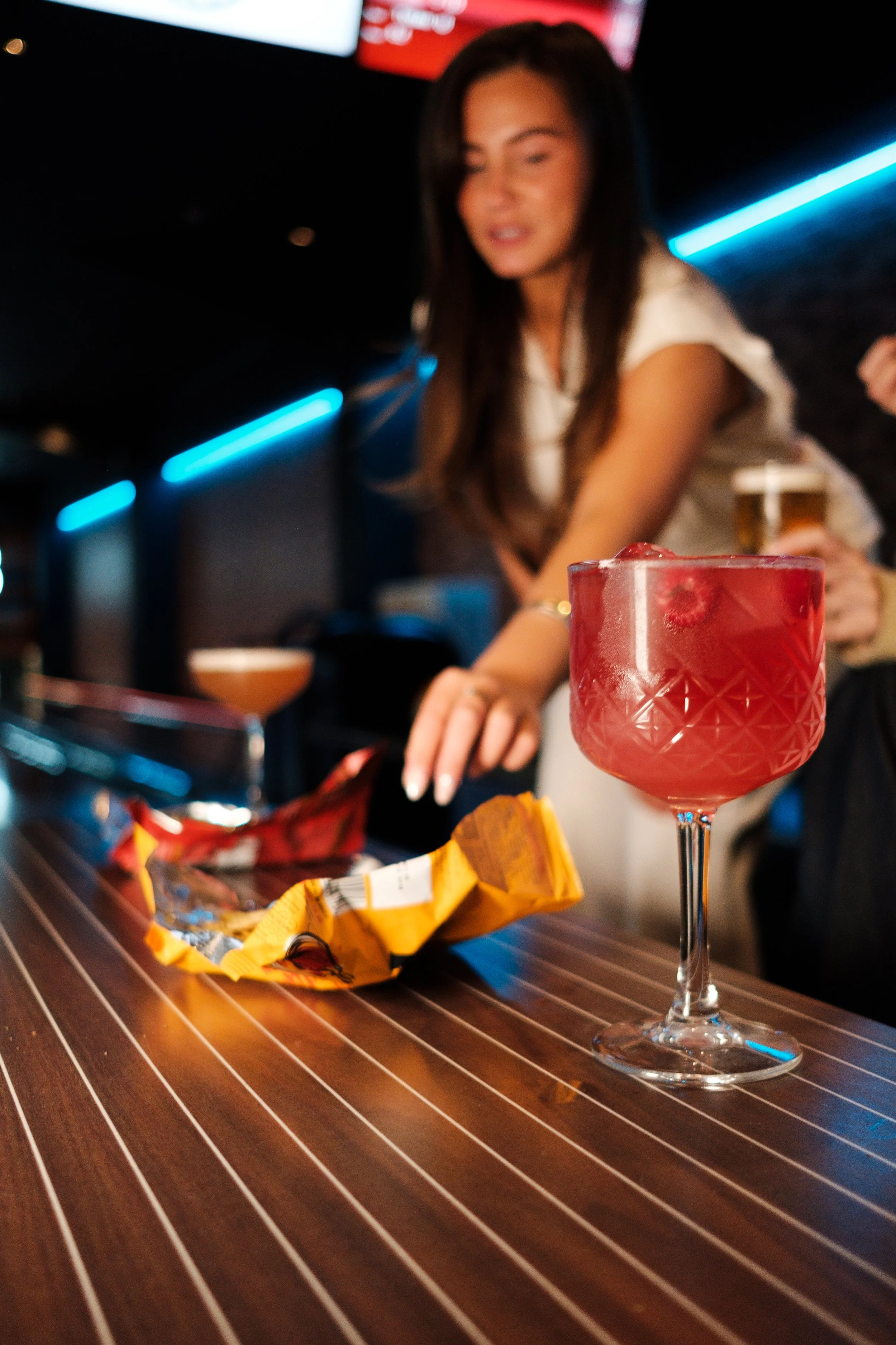 A woman with long brown hair in a white top leaning over a bar in a dimly lit setting, with a pink cocktail with a frosted rim and a yellow snack bag on a striped wooden table in the foreground.