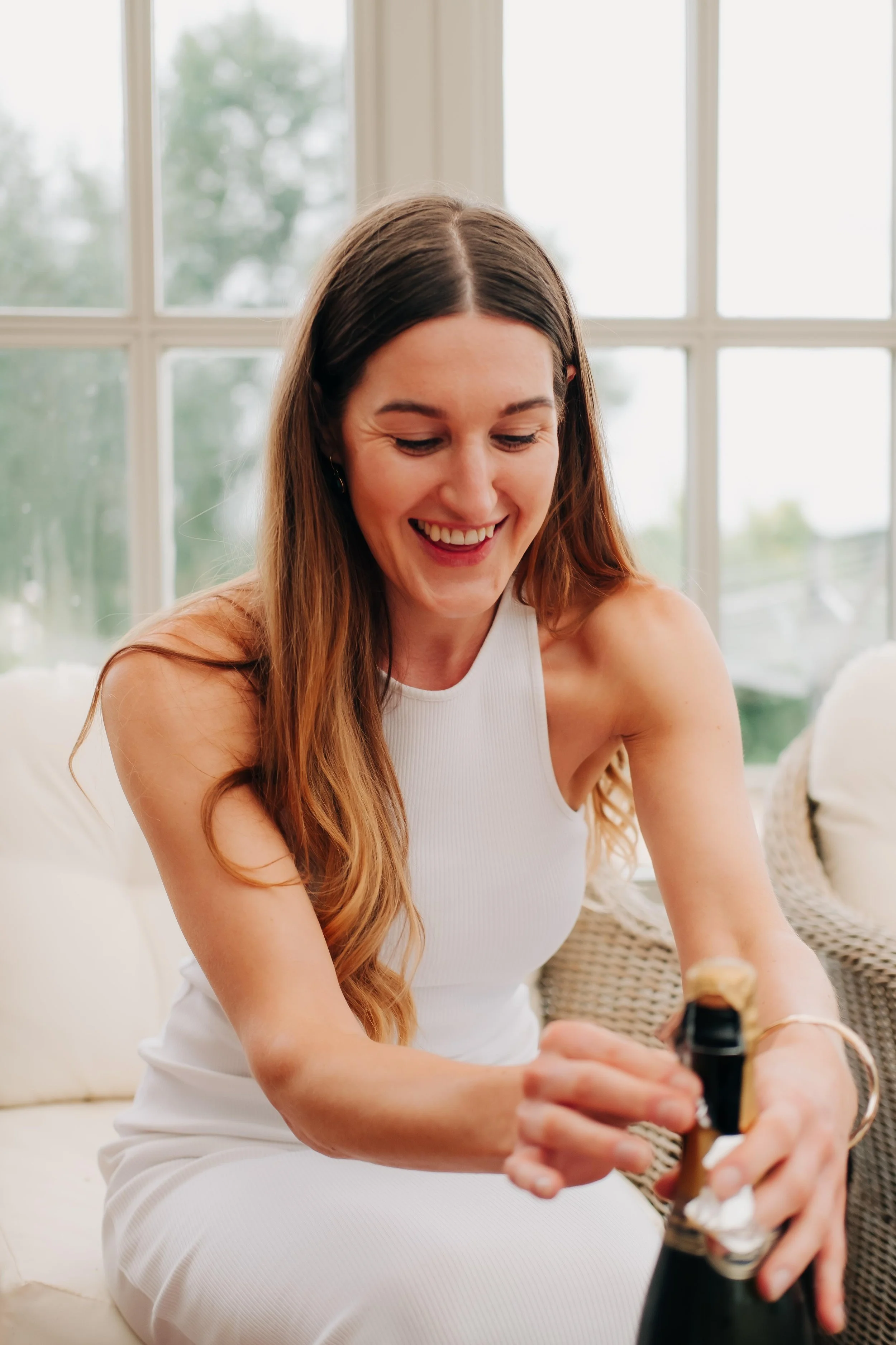 A woman with long brown hair in a white sleeveless top is smiling and looking down while opening a bottle of champagne. She is sitting indoors in front of a large window with a blurred outdoor view.