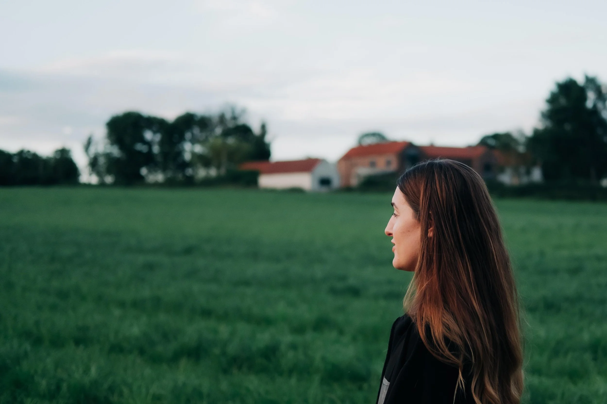 A woman with long brown hair standing outdoors in a grassy field with houses and trees in the background.