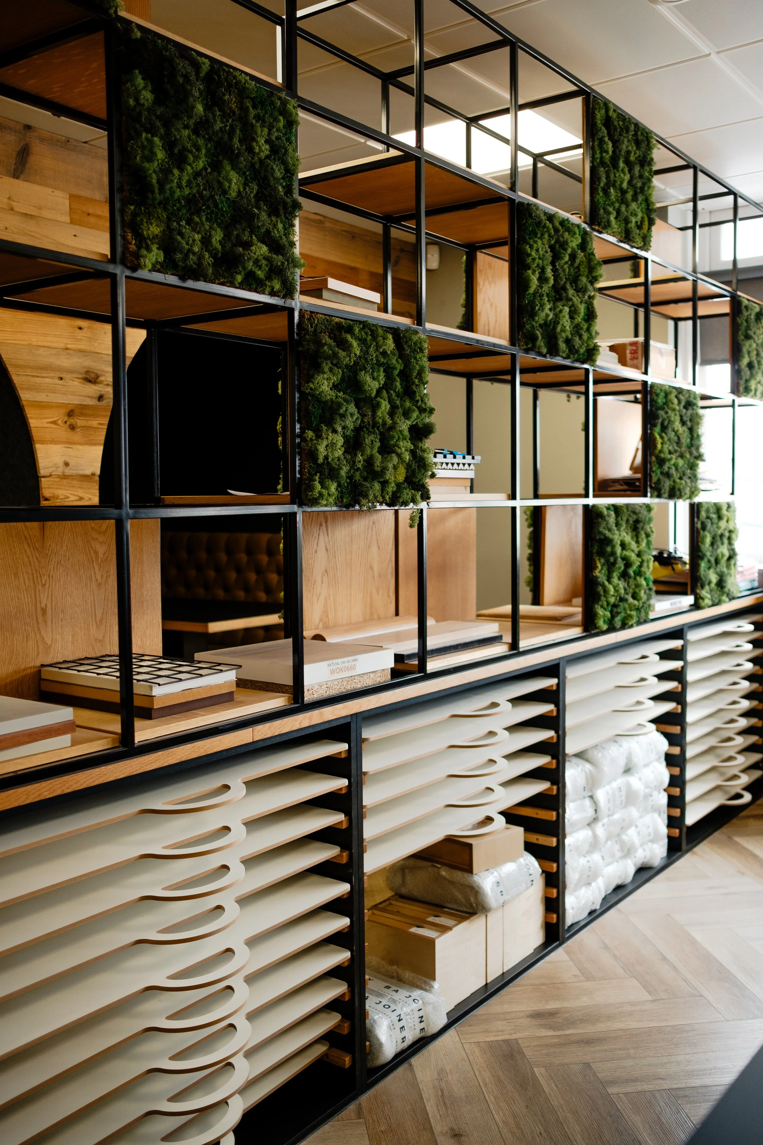 A modern shelving unit with black metal frame and wooden shelves, containing land and interior design samples, green moss decorative panels, and storage boxes, in a showroom or store.