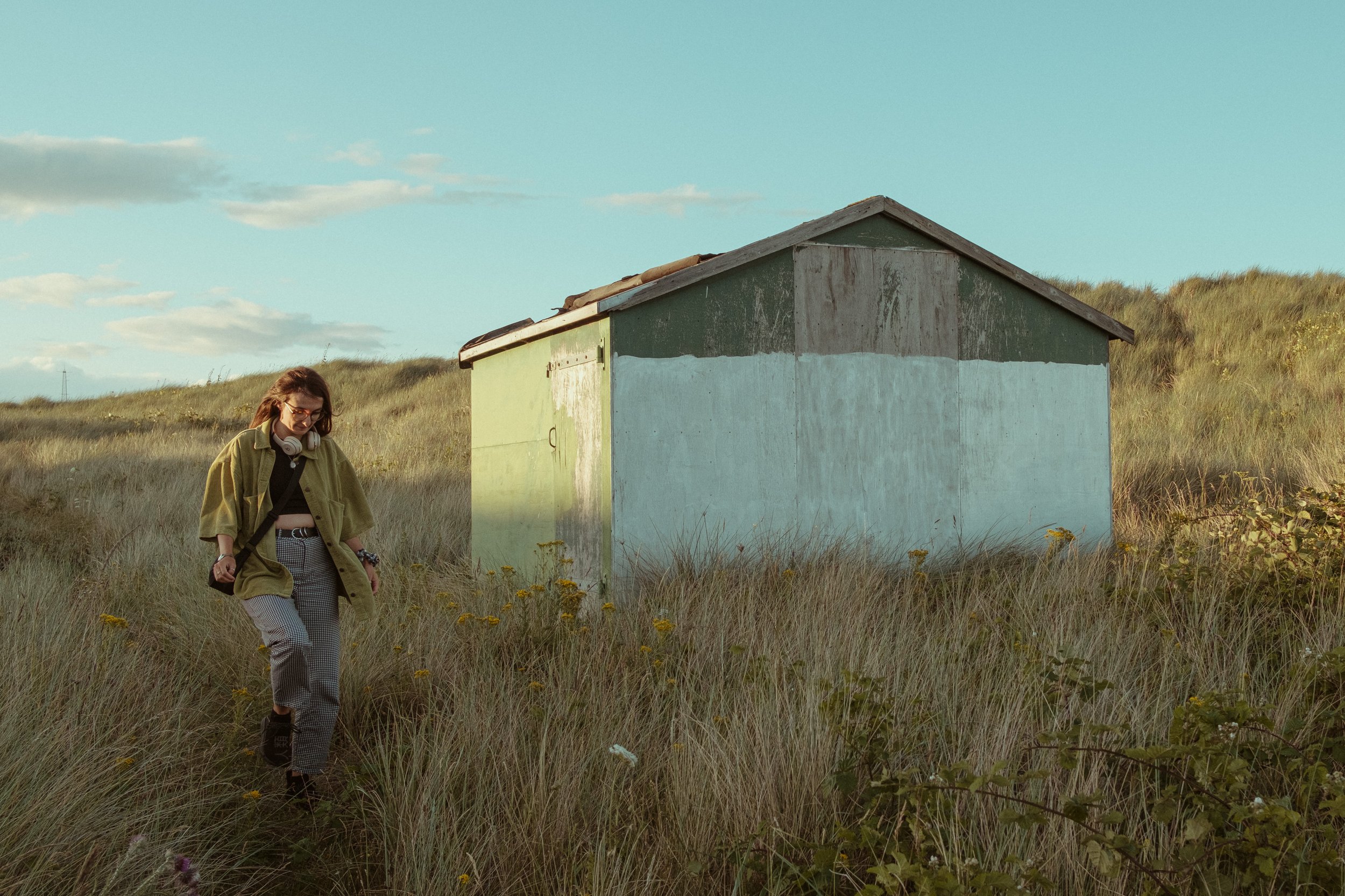 A woman walking through grassy field near a small, weathered green shed during daytime.