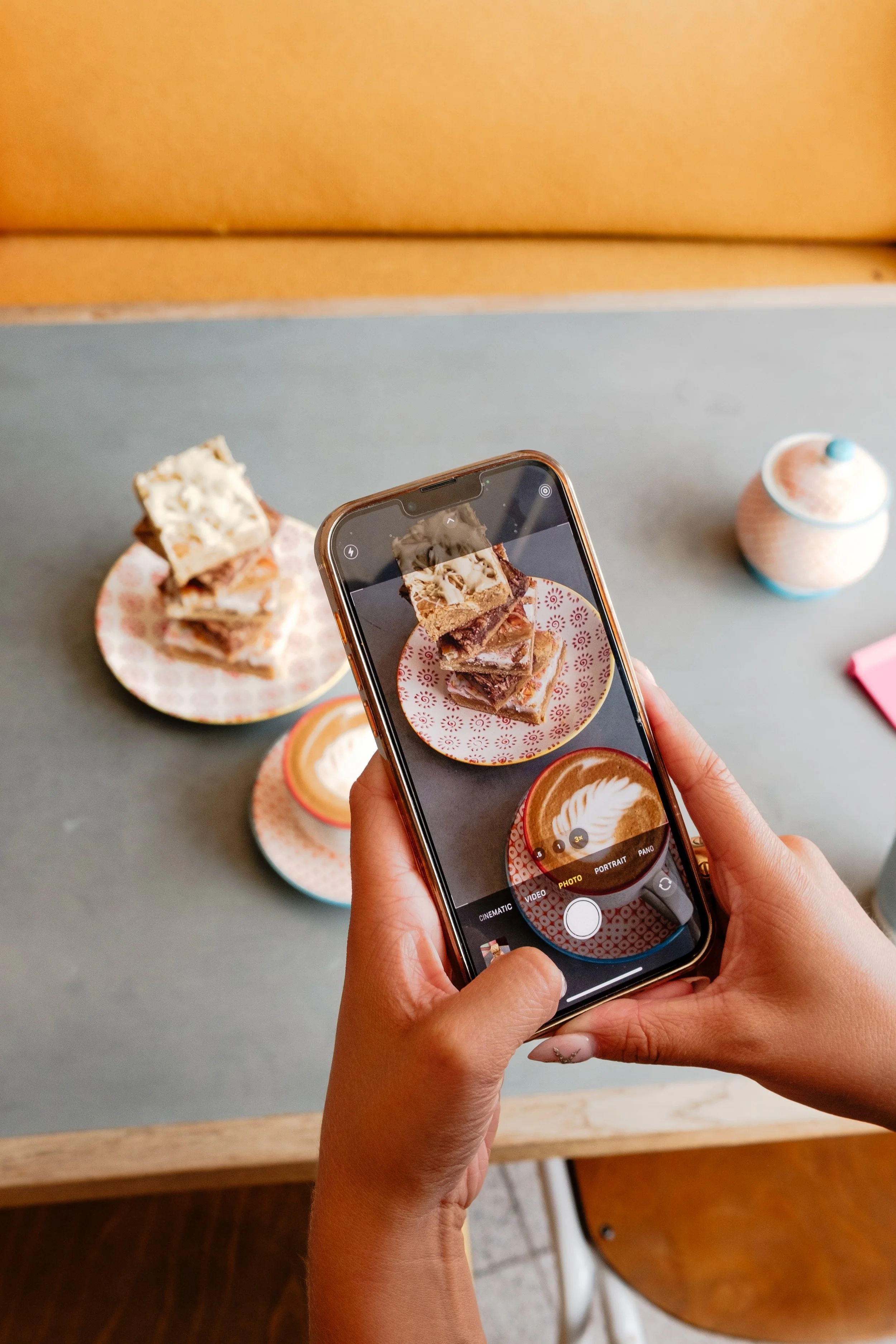 A person taking a photo of a plate of layered dessert and coffee drinks using a smartphone in a café.
