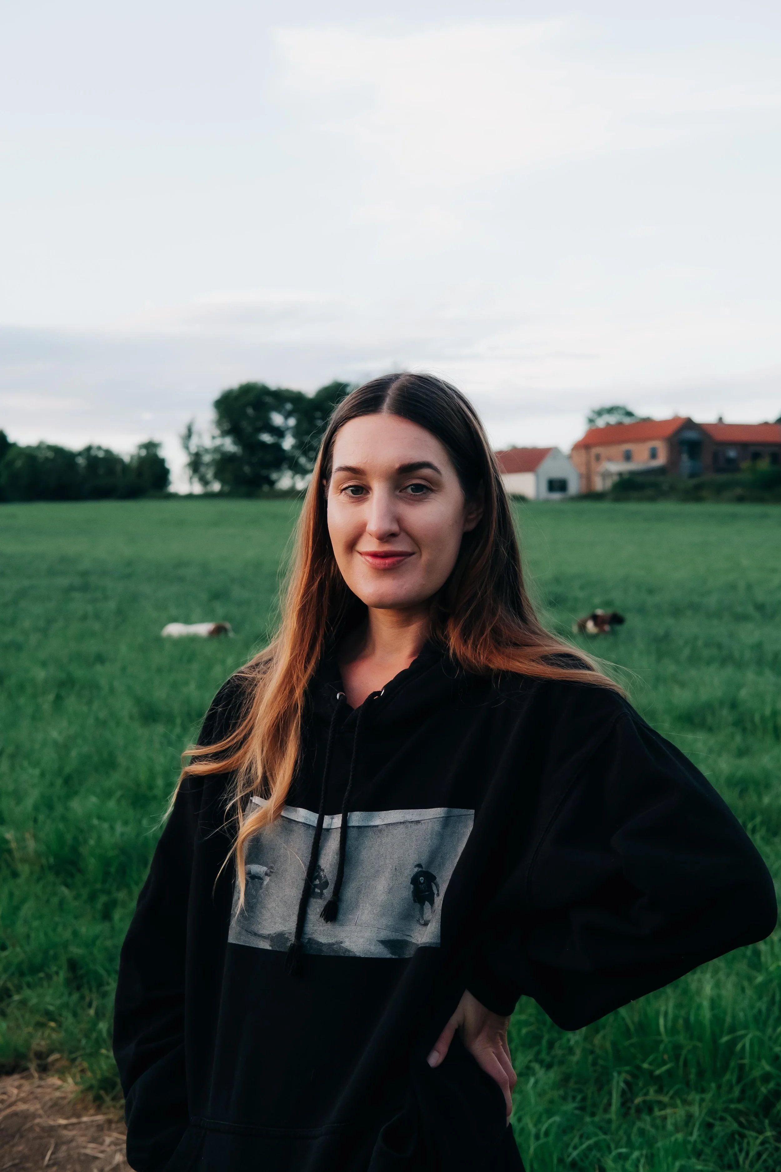 A young woman with long brown hair standing outdoors in a green field with trees and houses in the background, wearing a black hoodie with a graphic on the front, in a rural setting during daylight.