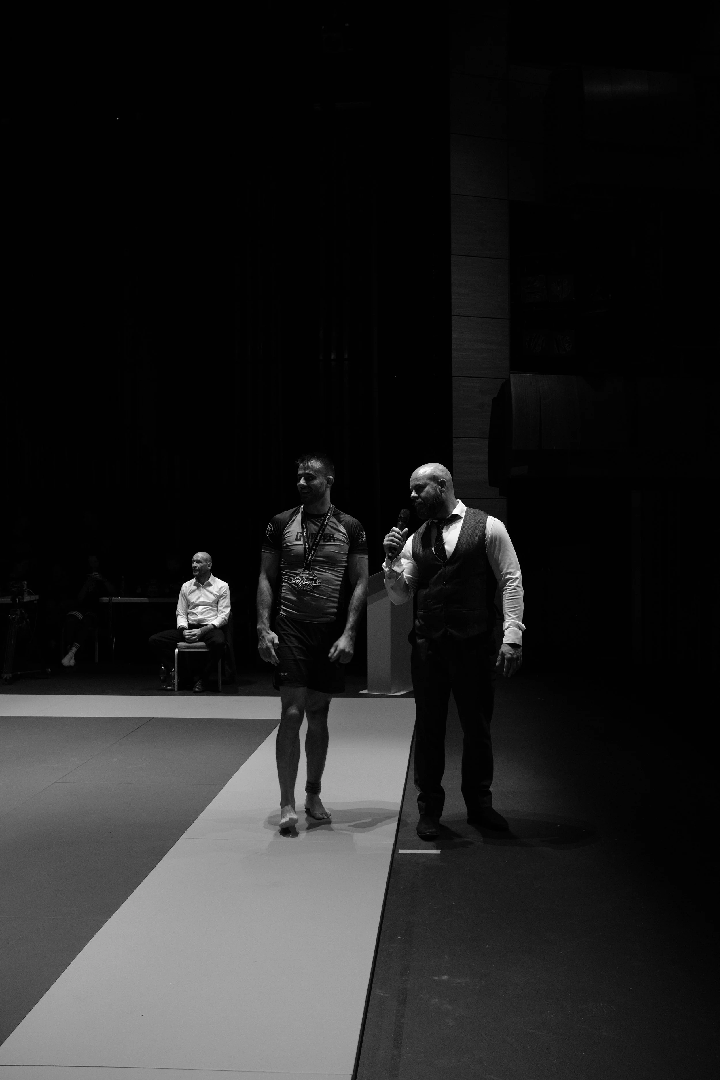 Black and white photo of a man in athletic clothing walking across a mat while another man in a vest and tie speaks into a microphone. Two other men are seated behind them in a darkened room.