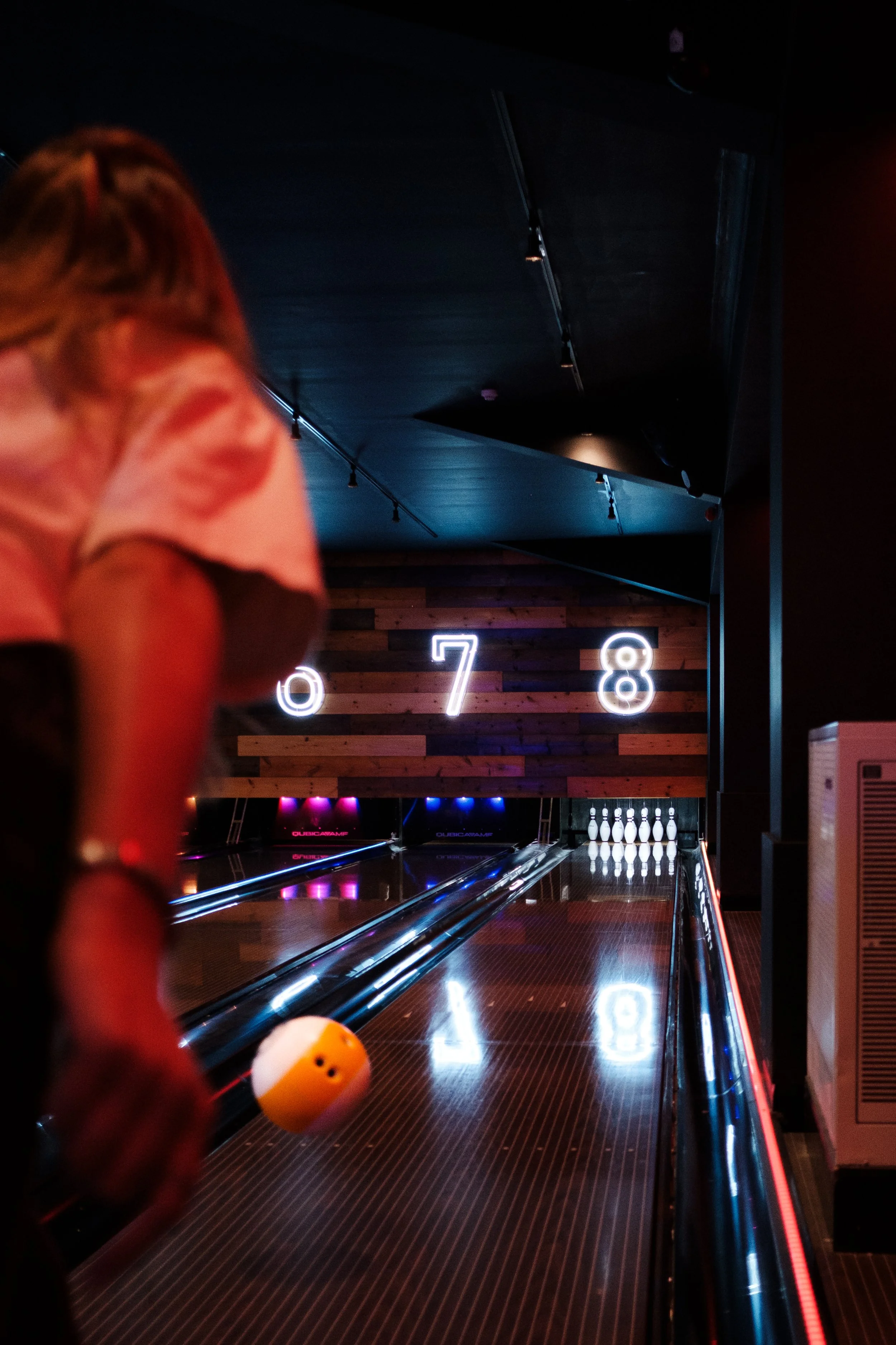 A person about to roll a bowling ball down a bowling lane, with digital score displays in the background.