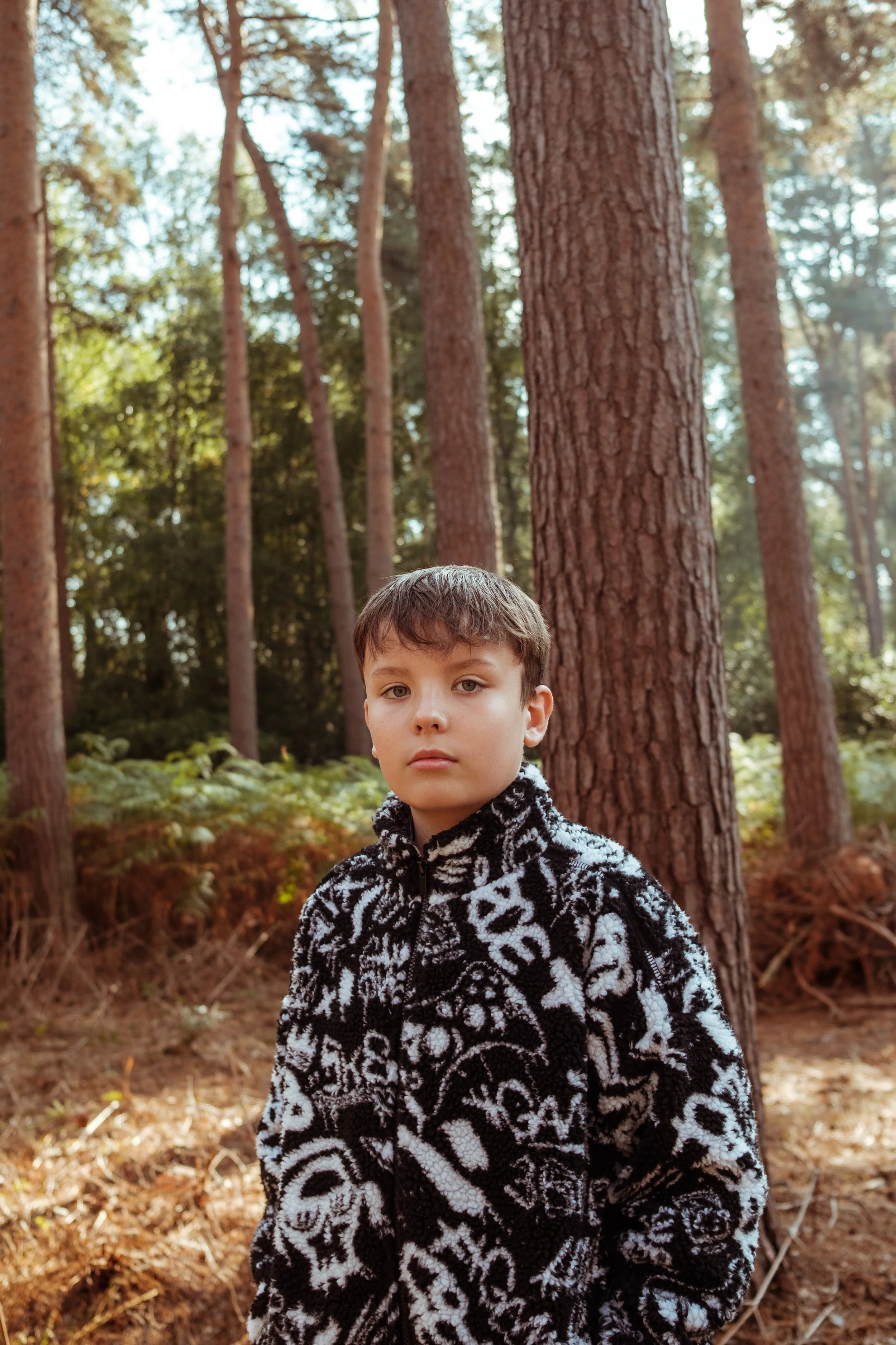A teenage boy with light brown hair and fair skin standing in a forest with tall trees and green foliage, wearing a black and white patterned fleece jacket.