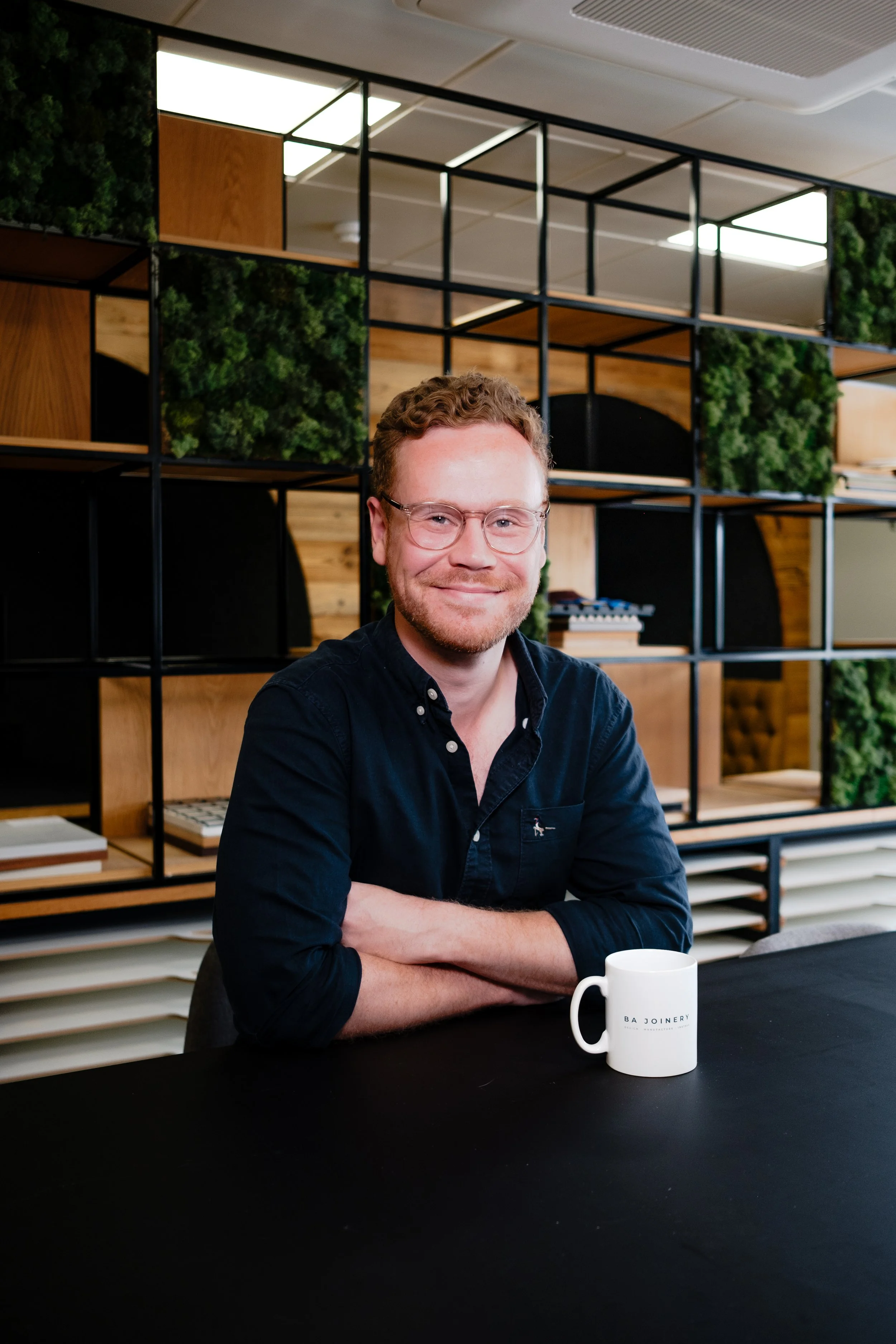 A man with glasses and a beard sitting at a table with a white mug, smiling at the camera in a modern office or coworking space, with a decorative wooden and green plant shelving unit behind him.