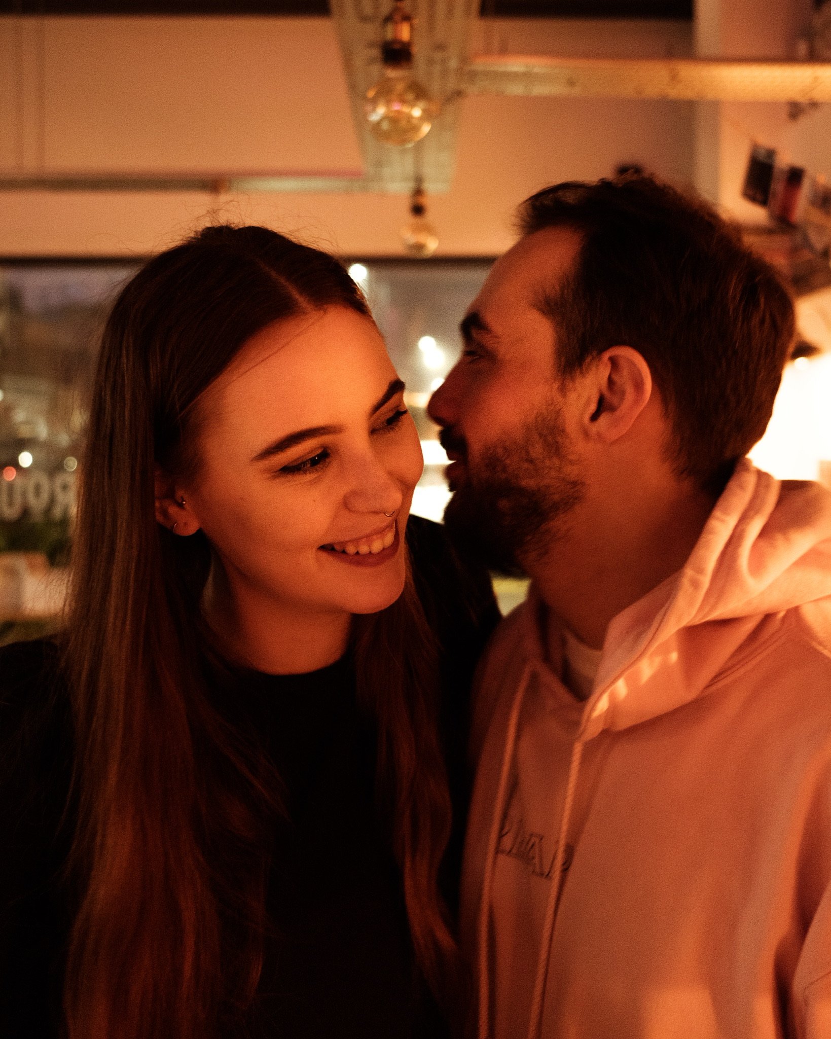 A close-up of a couple with light skin in an intimate moment, the woman smiling with her eyes closed and the man about to kiss her, in a warmly lit room.