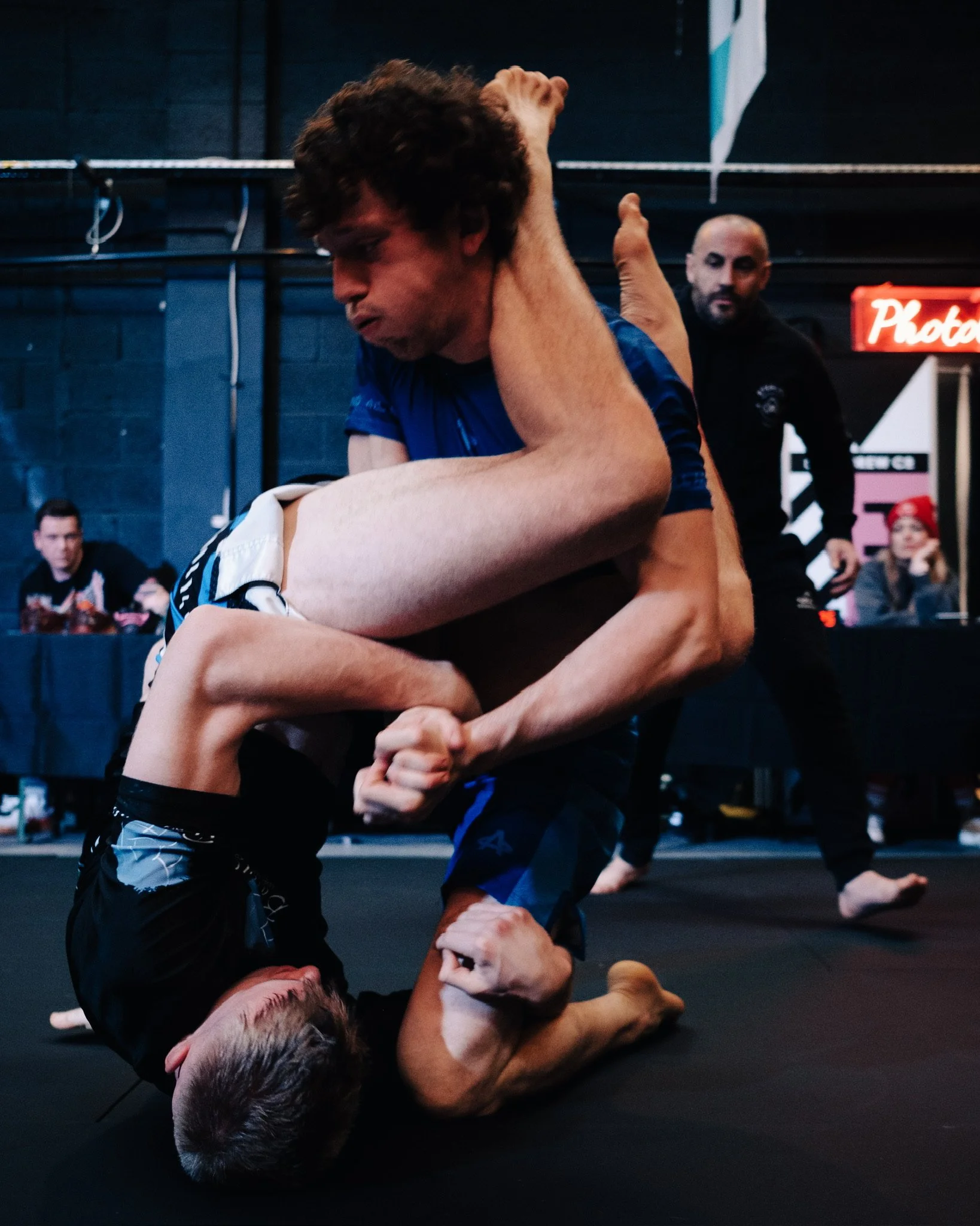 Two men competing in Brazilian Jiu-Jitsu grappling match on black mat, with spectators watching in background.