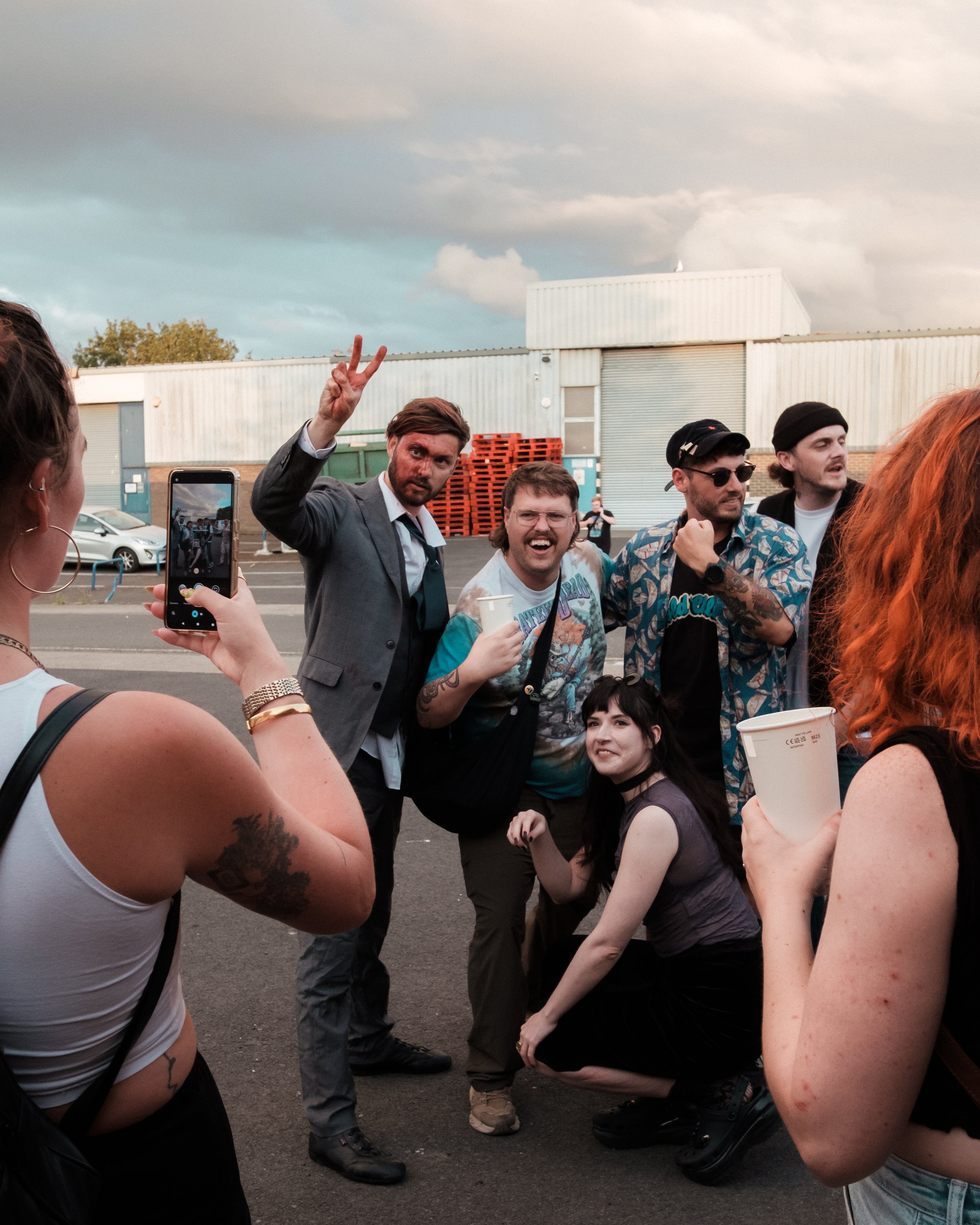 Group of young friends taking a photo together outdoors, some making peace signs and smiling, in a parking lot with a warehouse behind them.