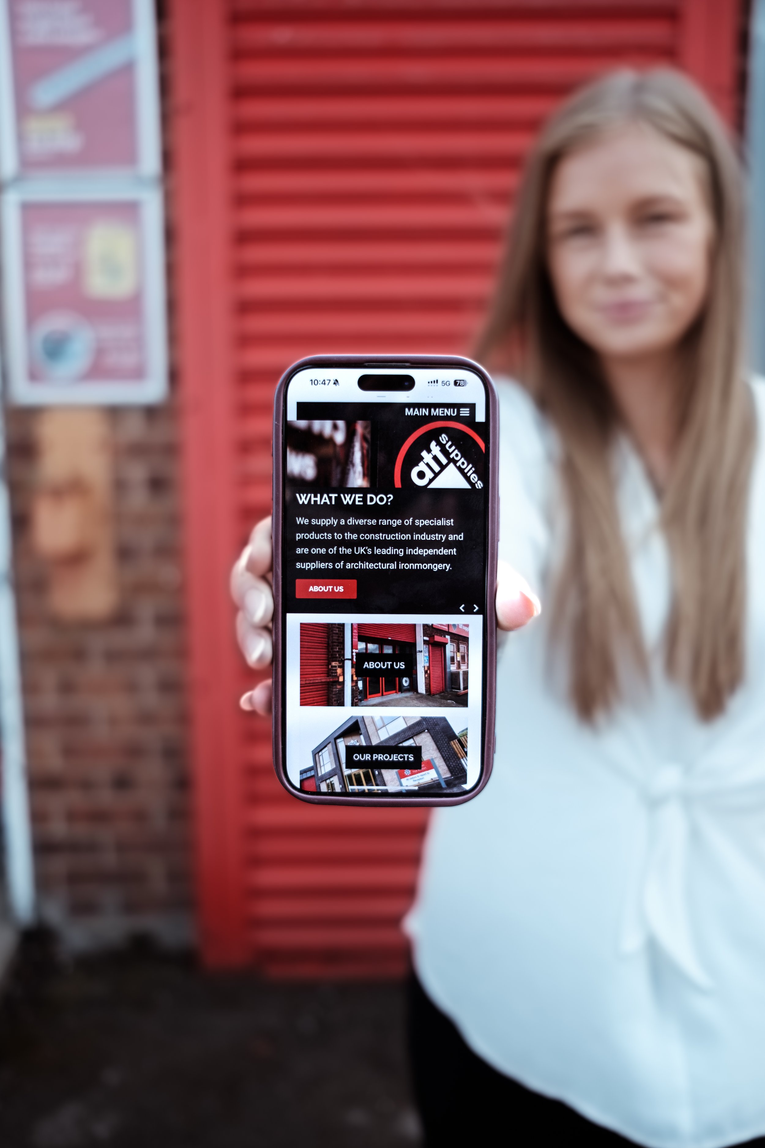 A woman holding a smartphone displaying a website for a company called 'Attrib Supplies,' with a background of a brick wall and a red paneled structure.