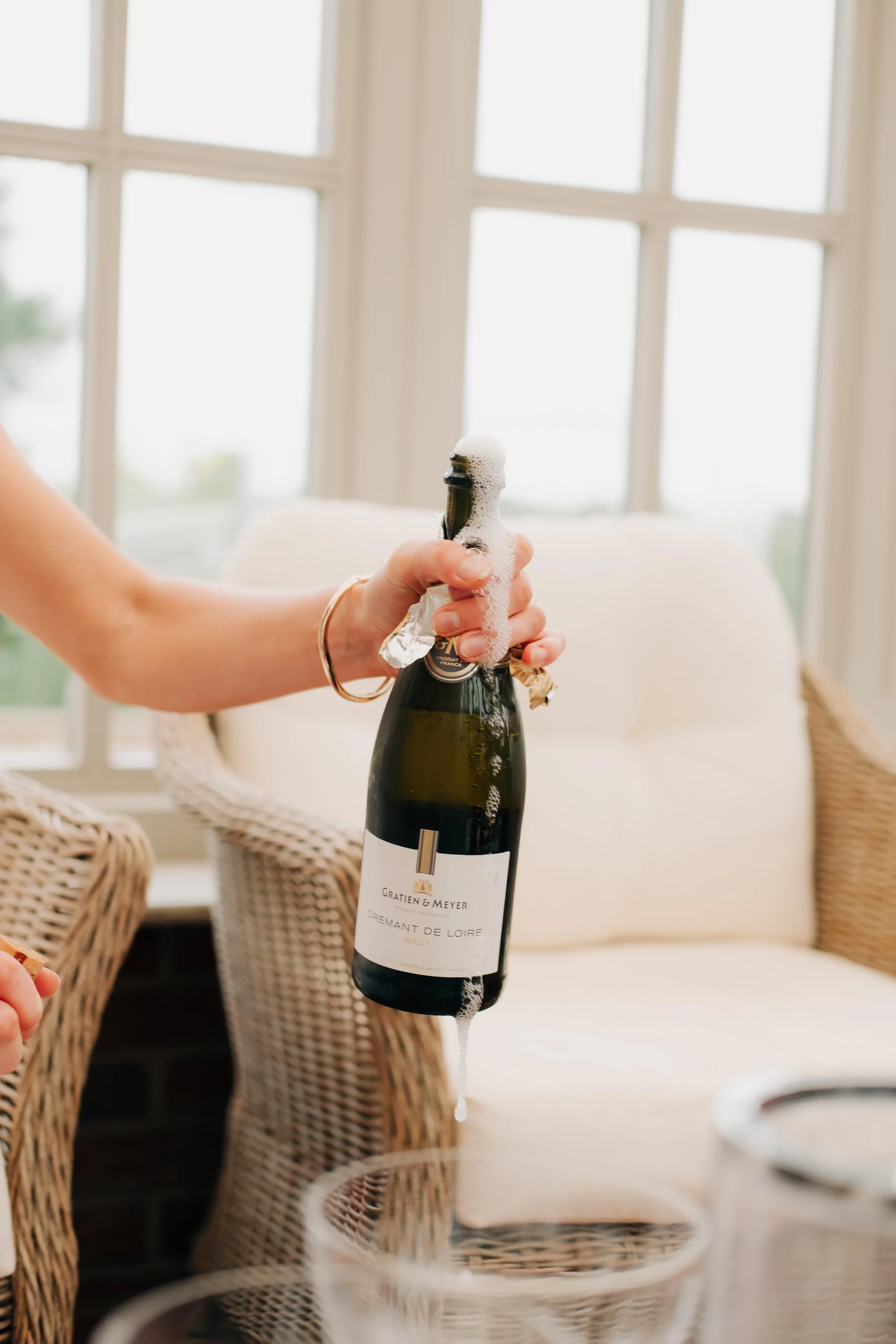 A person holding a champagne bottle with foam overflowing at a dining table near wicker chairs and a window.