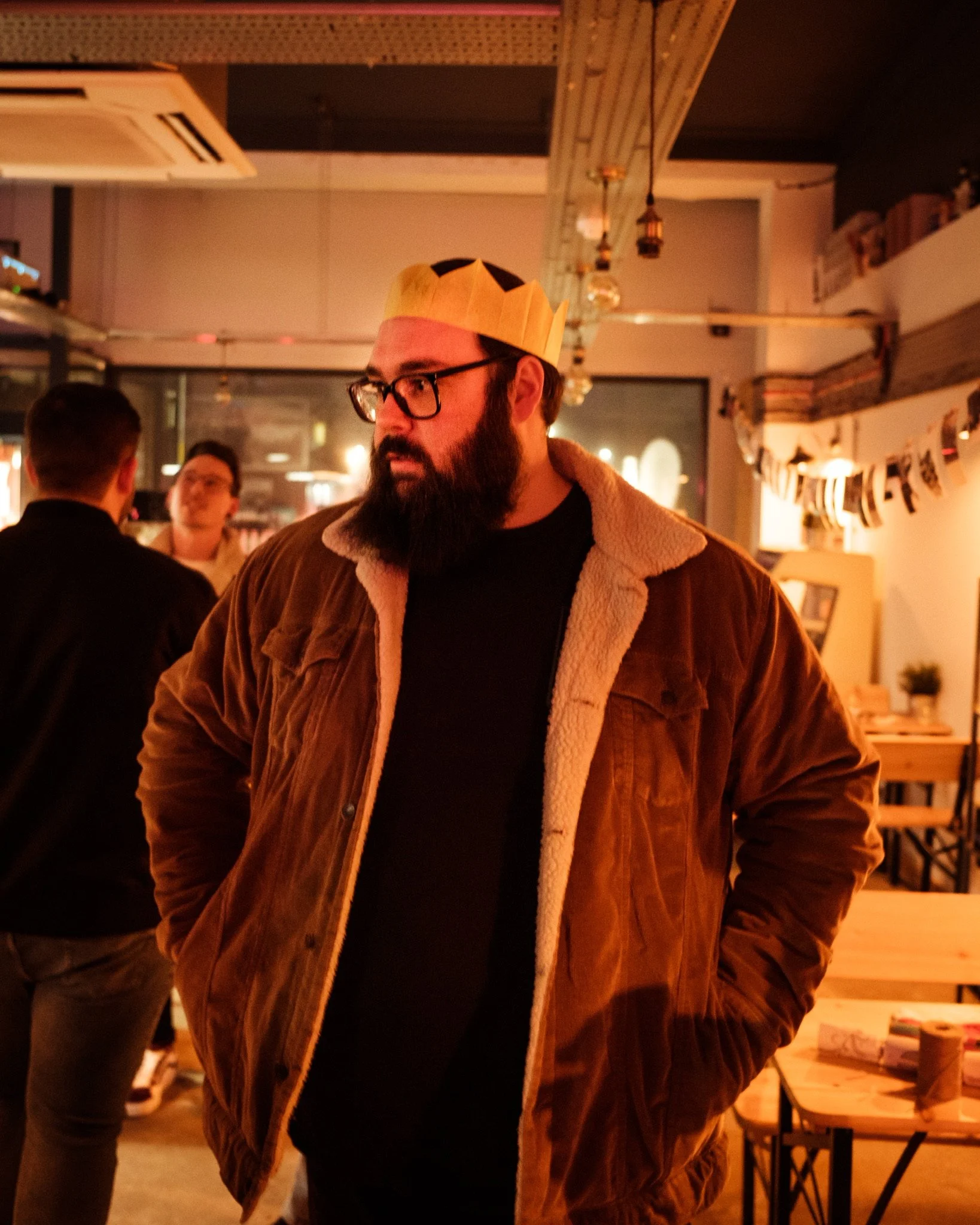 A man with glasses and a beard wearing a paper crown in a cozy, warmly lit cafe or restaurant. Other people are visible in the background, and festive decorations hang from the ceiling.