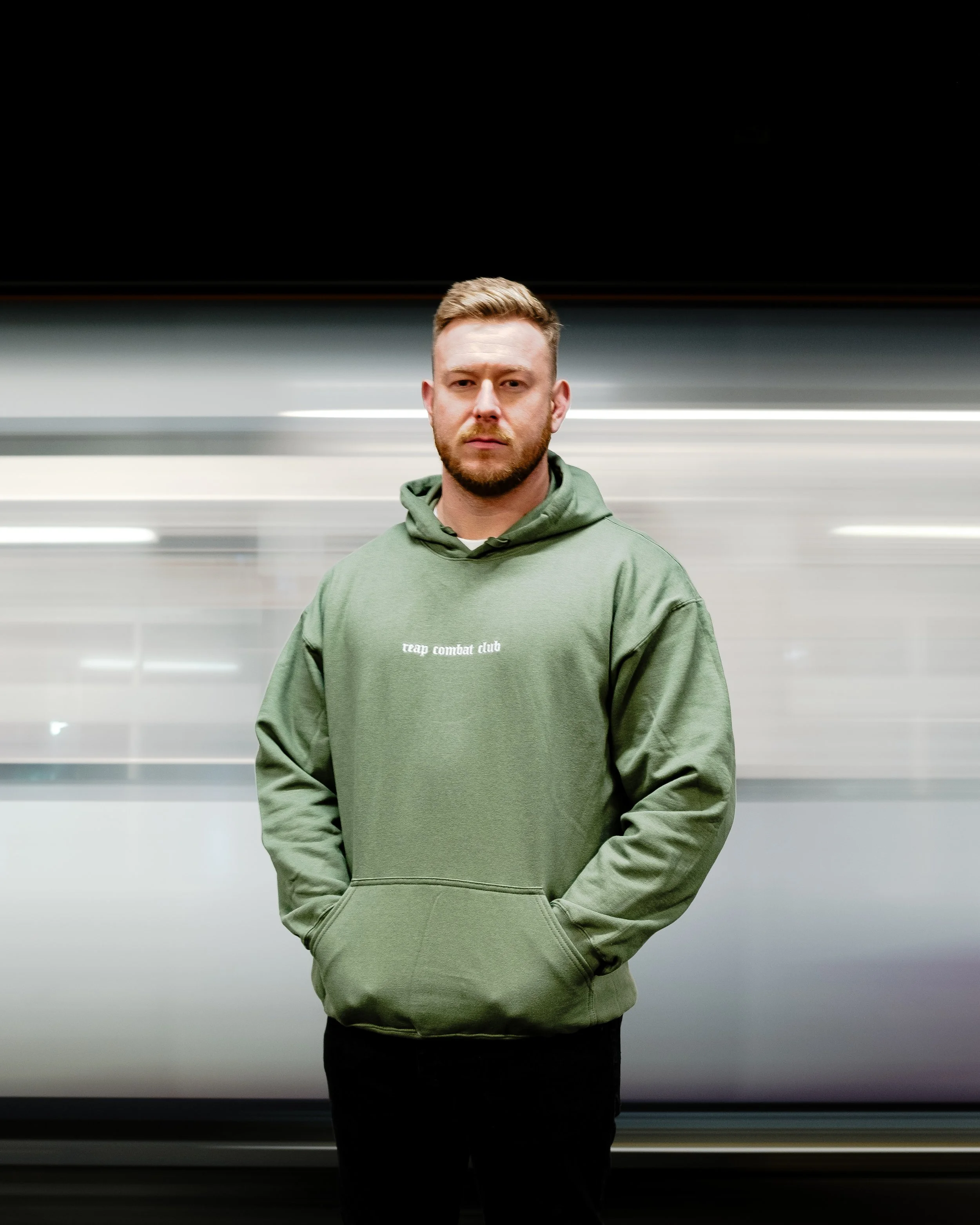 A man in a green hoodie stands in front of a blurred background, looking directly at the camera with a serious expression.