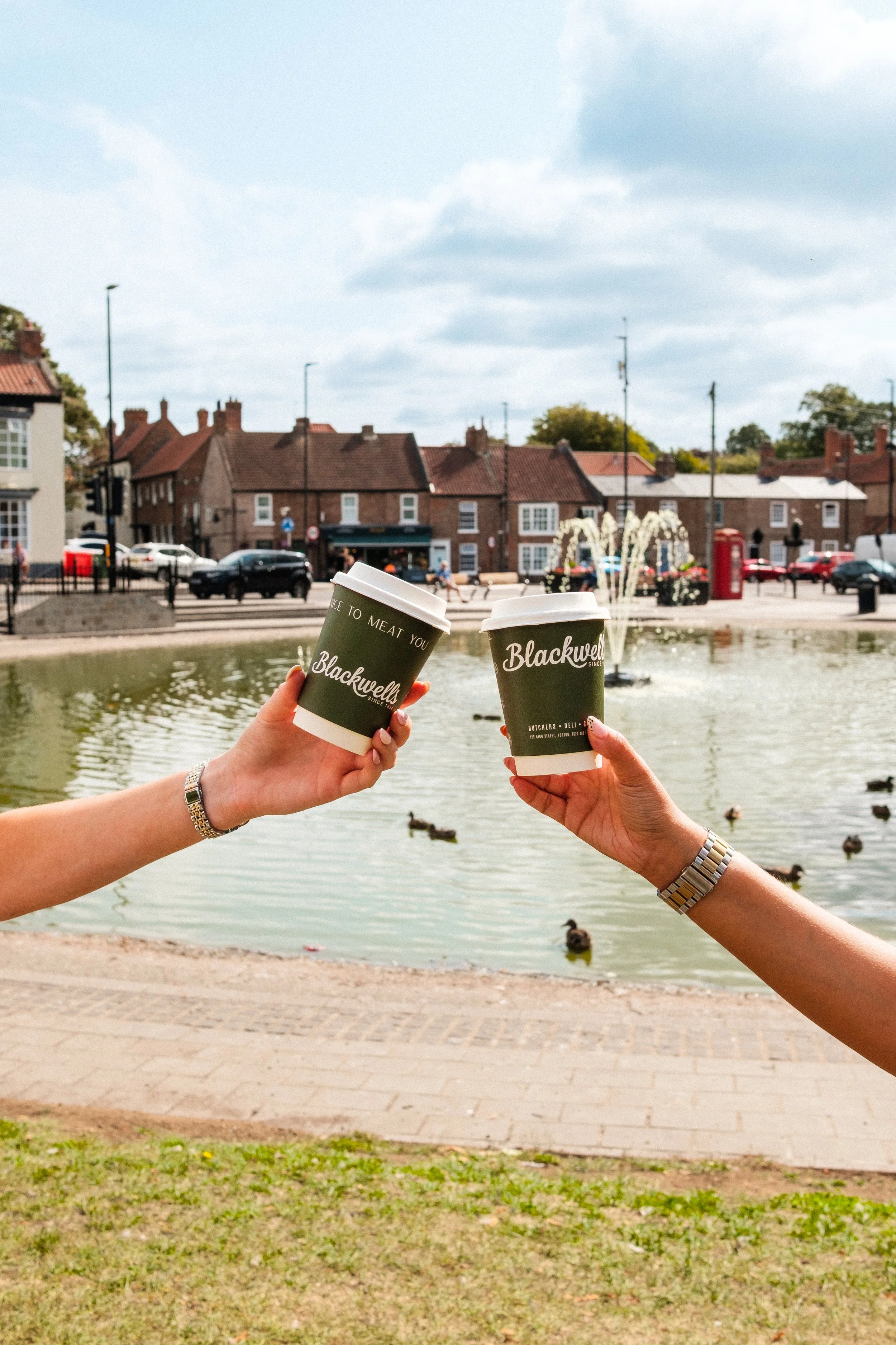 Two hands holding coffee cups with a town square, a pond with ducks, and old brick buildings in the background on a cloudy day.