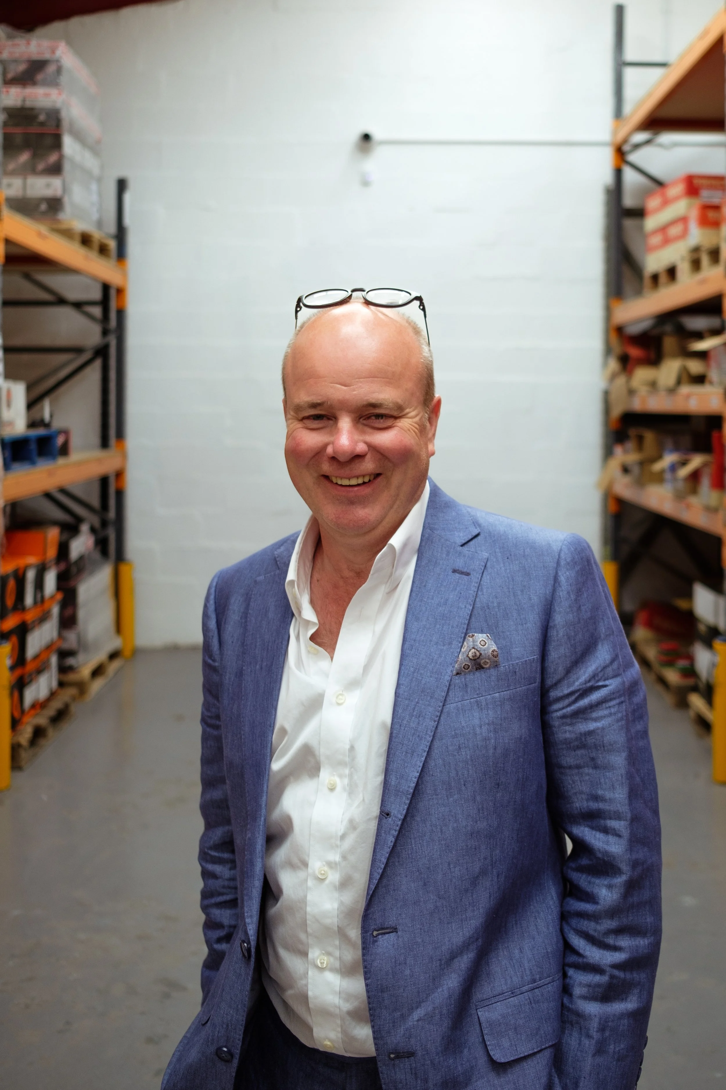 A smiling man in a blue suit jacket and white shirt standing in a warehouse with orange shelving filled with boxes on both sides.