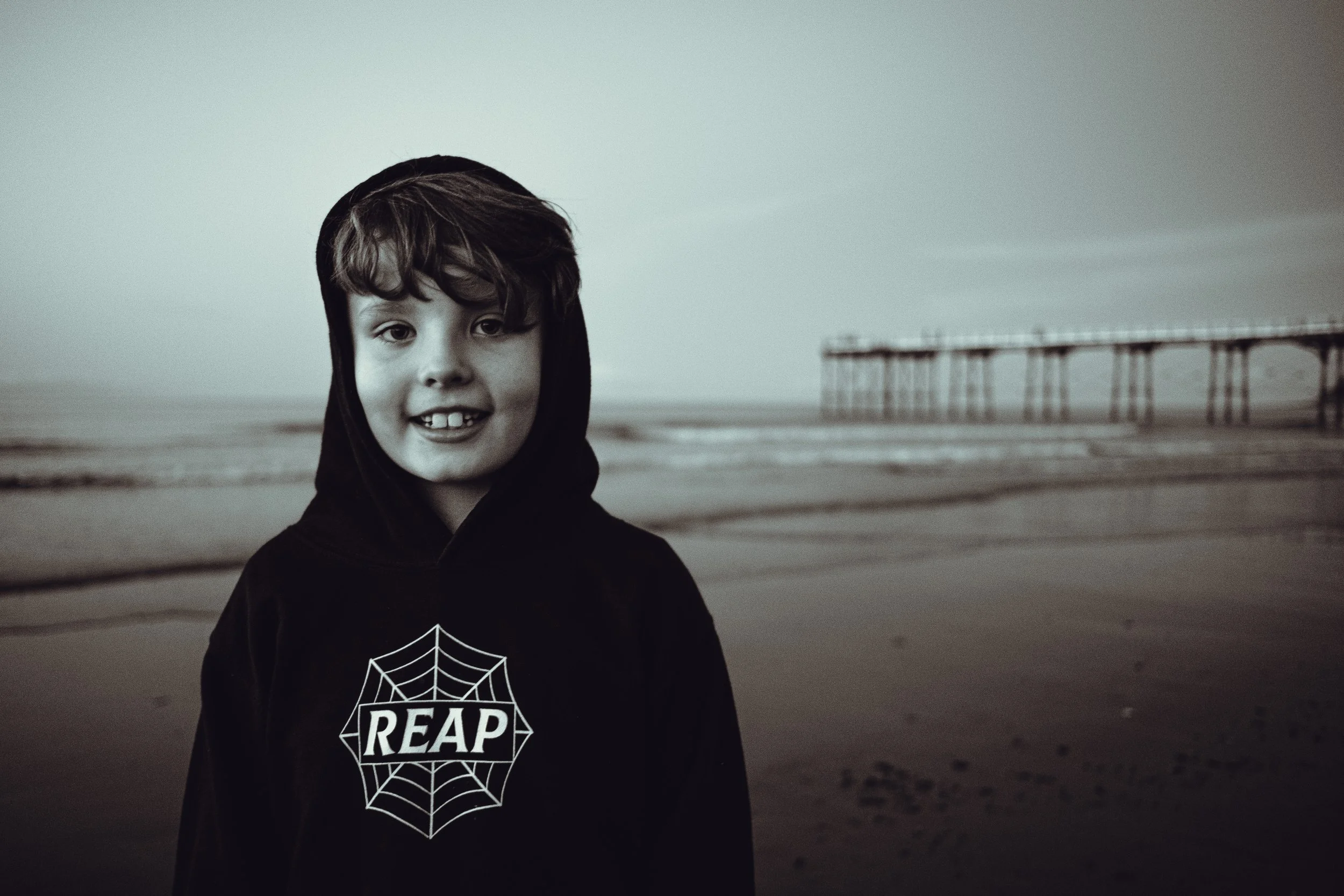 A young boy with dark hair smiling at the camera, standing on a beach with a pier in the background, dressed in a black hoodie with a spider web and the word REAPER on it.