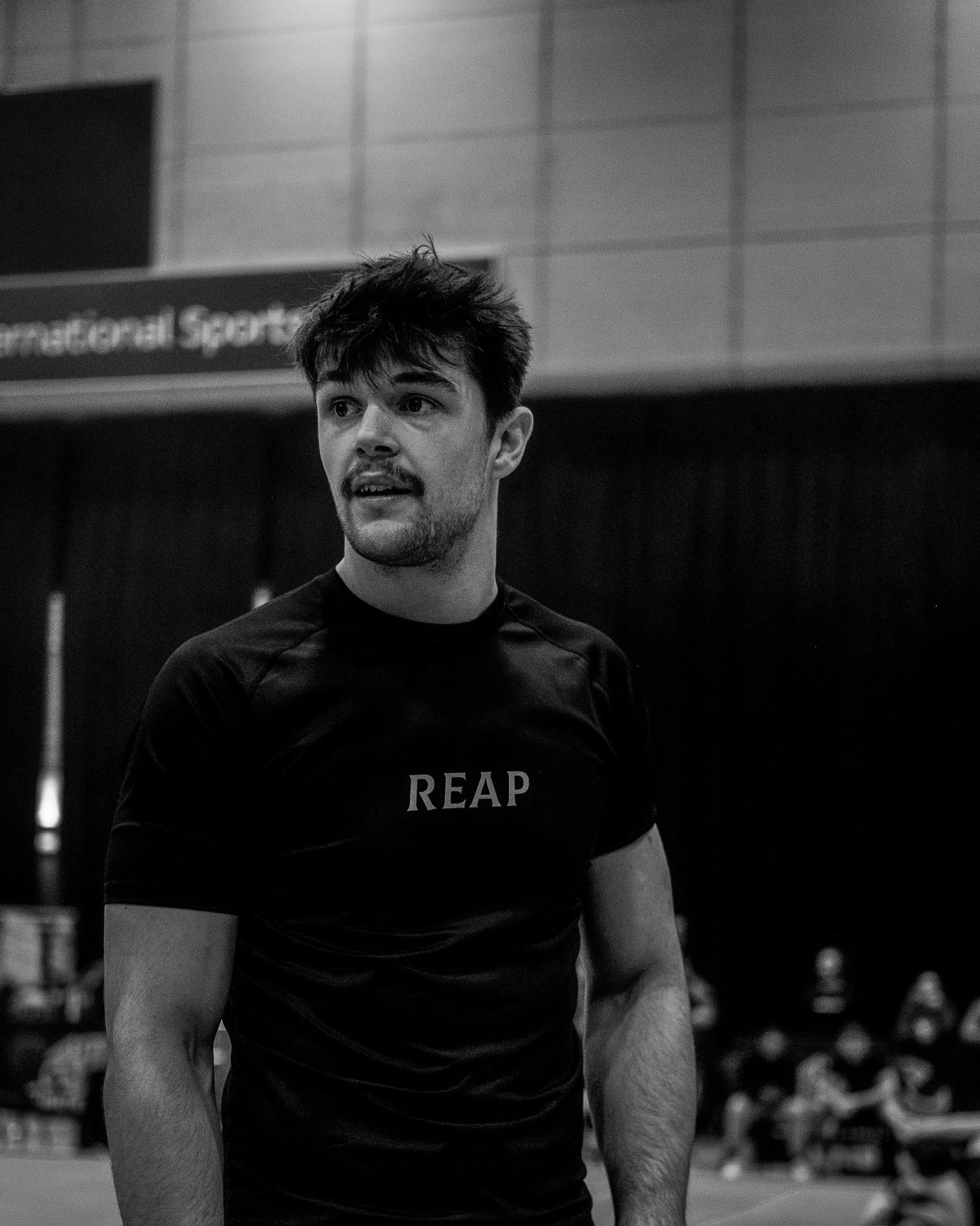 A young man with dark hair and a beard, wearing a black REAP T-shirt, standing indoors in a sports facility, likely a gym or gymnasium.