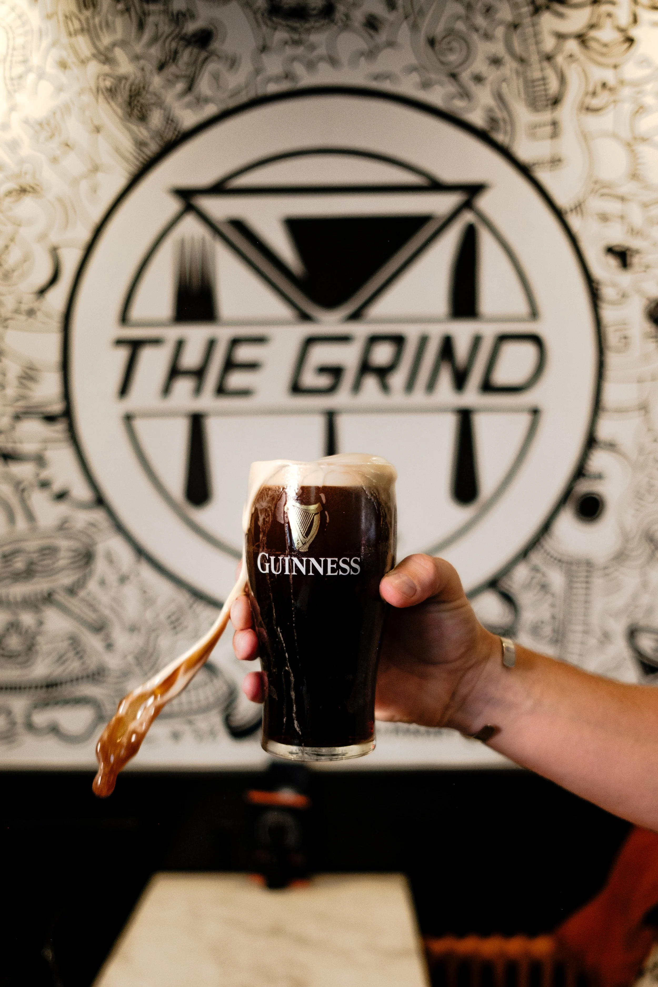A hand holding a Guinness pint with foam overflowing, in front of a wall with a sign that reads 'The Grind'