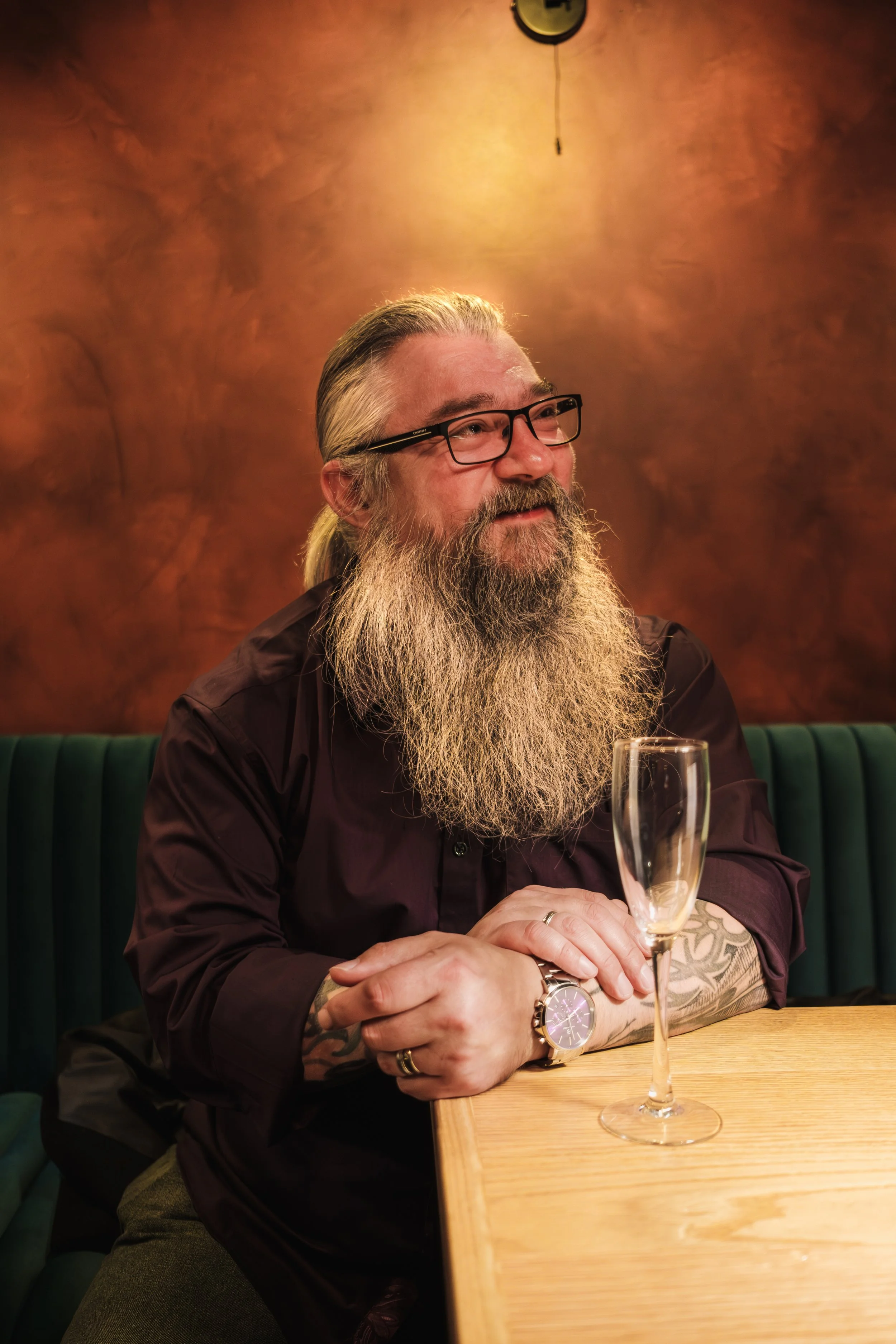 A man with a long gray beard, glasses, and tattoos on his arms sitting at a wooden table with a glass of champagne in front of him. The background has warm-colored walls and a clock hanging on the wall.