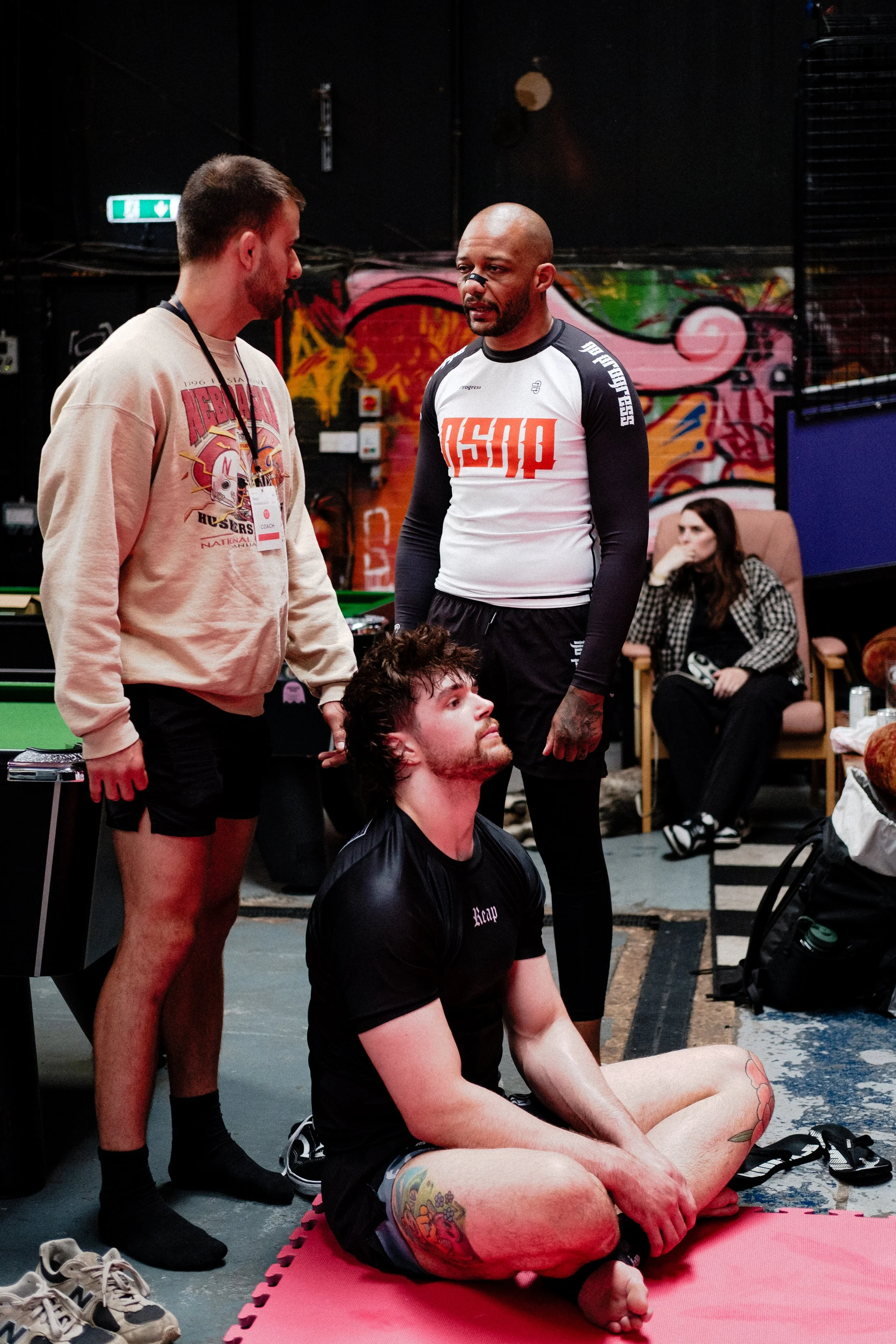 Two men with athletic builds and tattoos, one sitting cross-legged on a pink mat and the other standing, are having a conversation in a gym. The background features a colorful graffiti wall, a woman sitting in a chair watching, and gym equipment.