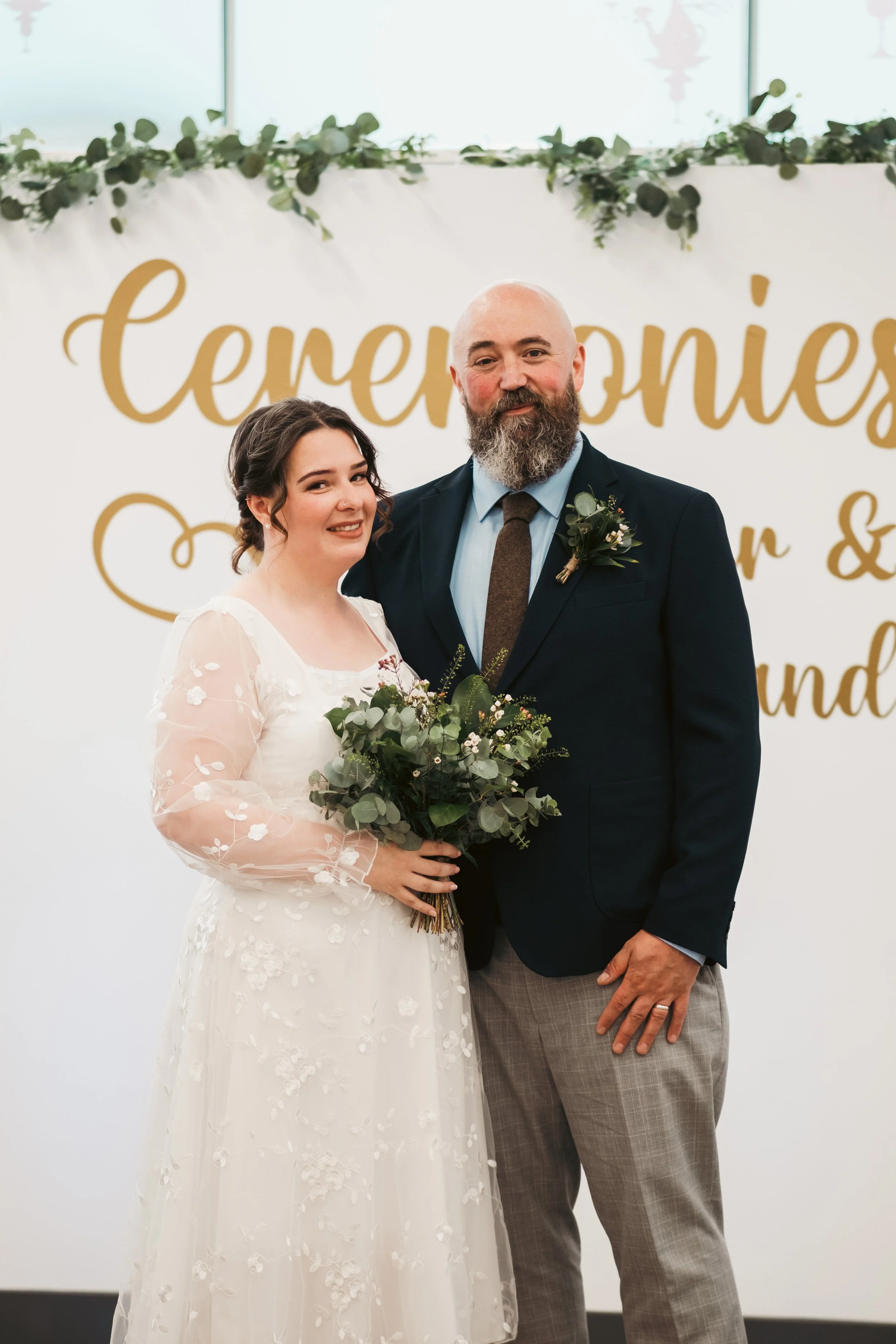 A bride and groom smiling at an indoor wedding ceremony, standing in front of a blurred sign that reads 'Ceremonies' with decorative greenery at the top.