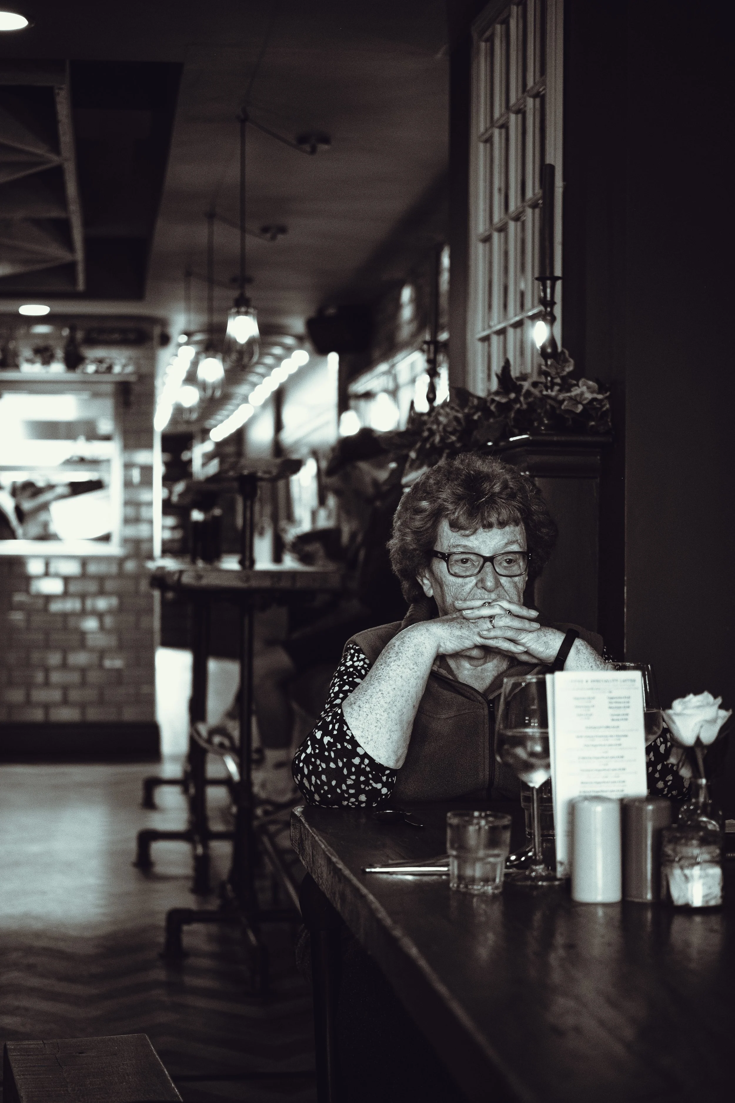 An elderly woman with glasses sitting alone at a restaurant table, resting her chin on her hands, looking pensive.