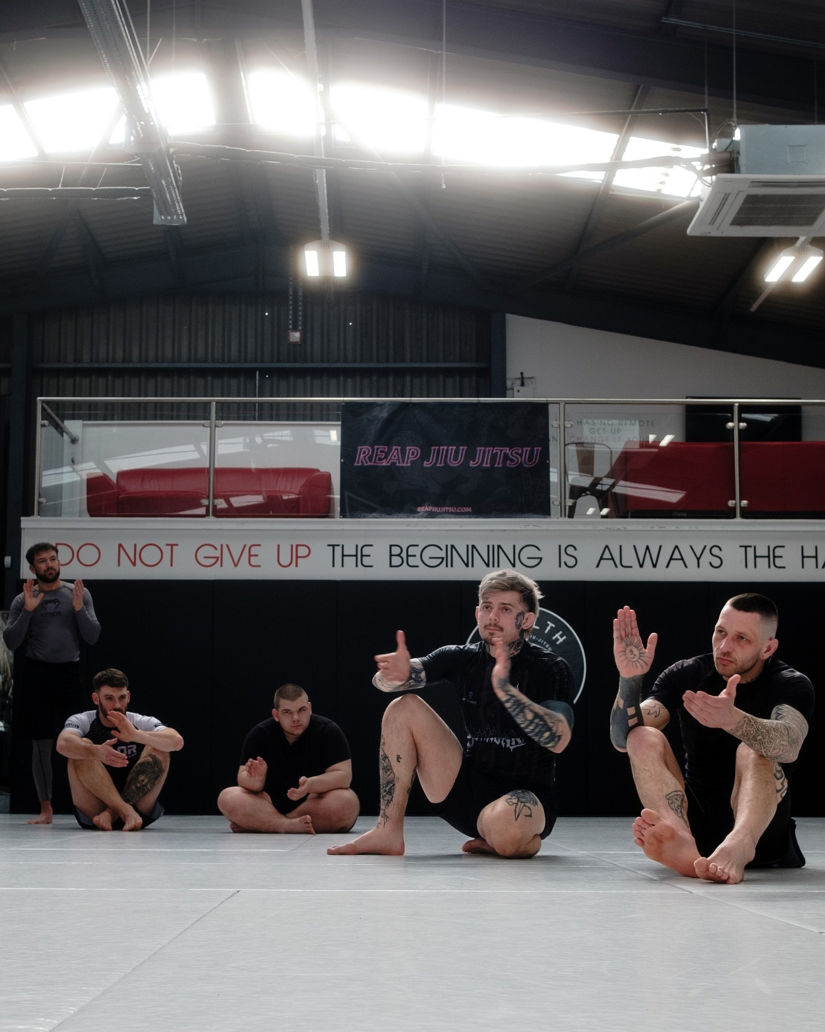 Martial arts students sitting on mats in a gym, listening to instructors, with a banner showing 'Reap Jiu Jitsu' and a motivational quote about not giving up.